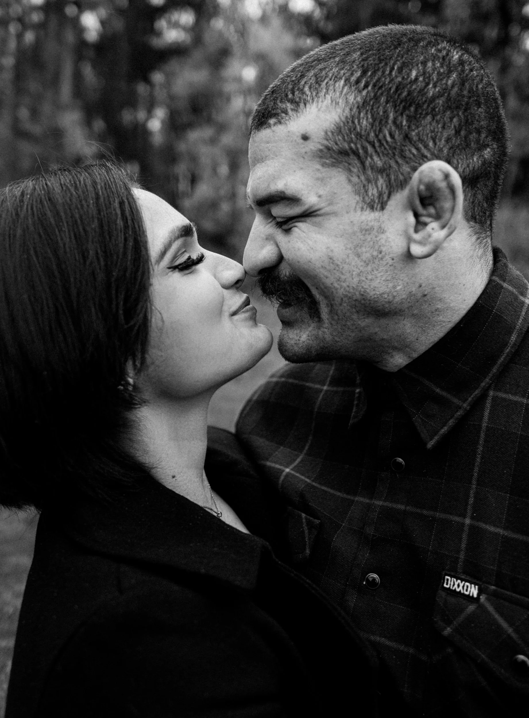 A black and white close-up photograph of a man and woman facing each other with their noses touching. They are smiling and appear to be in an outdoor setting with blurred background trees.