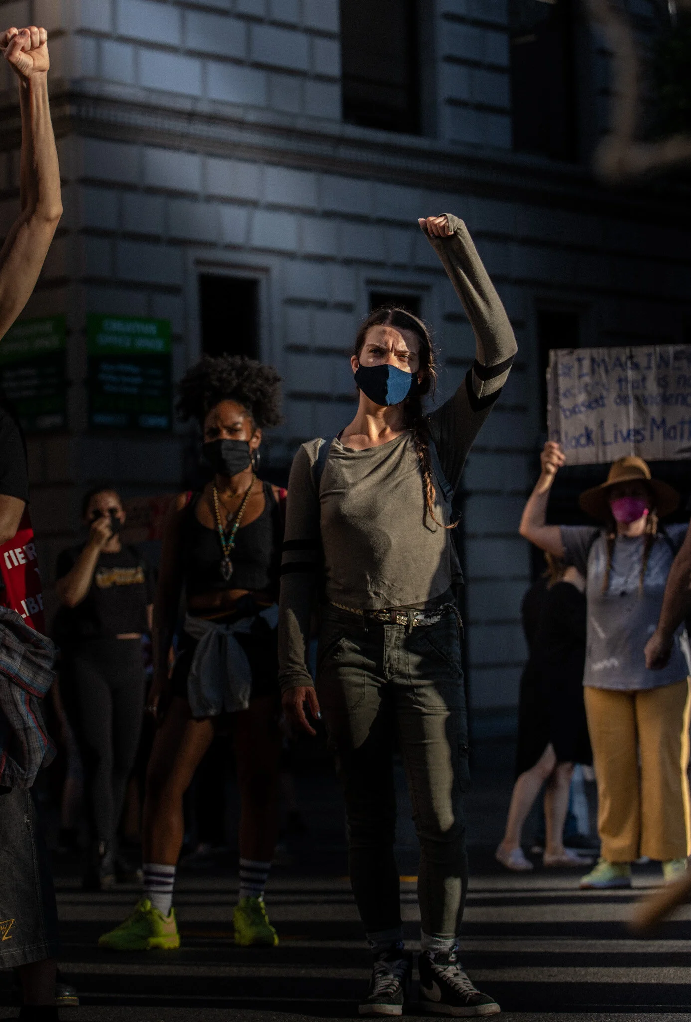 A group of people participating in a nighttime protest or demonstration on street steps, some wearing face masks, holding signs, with a woman in the center raising her fist.