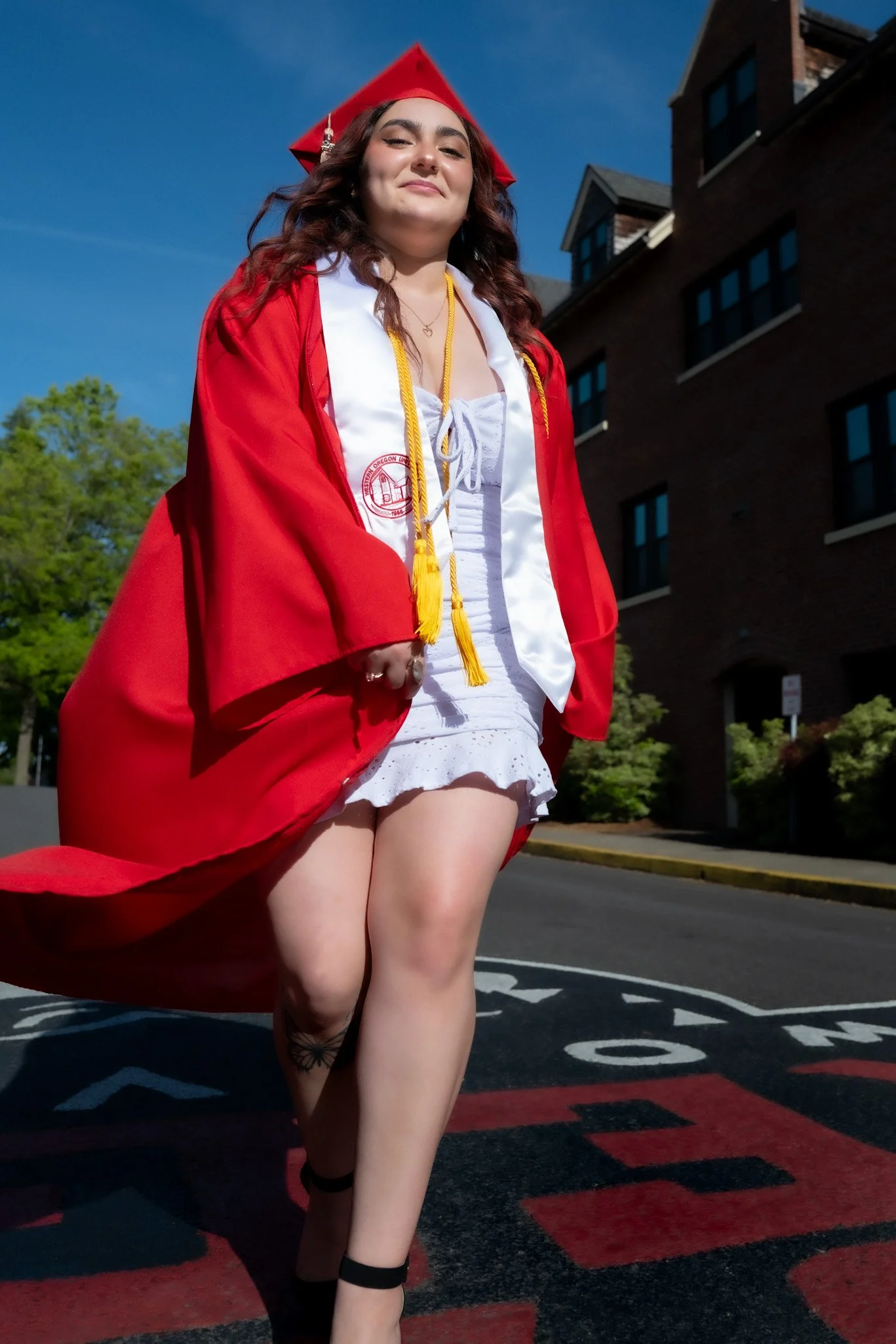 A young woman in a white dress and red graduation gown and cap with gold honor cords, standing confidently outside on a sunny day, smiling with her eyes closed, in front of a brick building and trees.