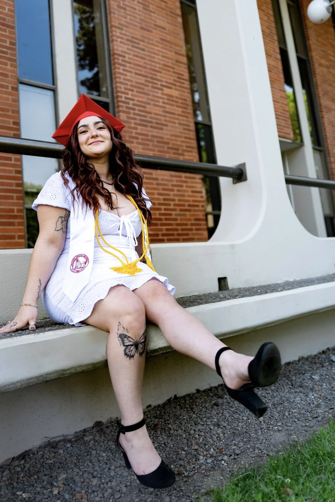 A young woman with long curly hair wearing a white dress, a red graduation cap, and a yellow graduation cord, sitting on a low concrete wall outside a building with red brick walls and large windows. She is smiling, with one leg crossed over the othe