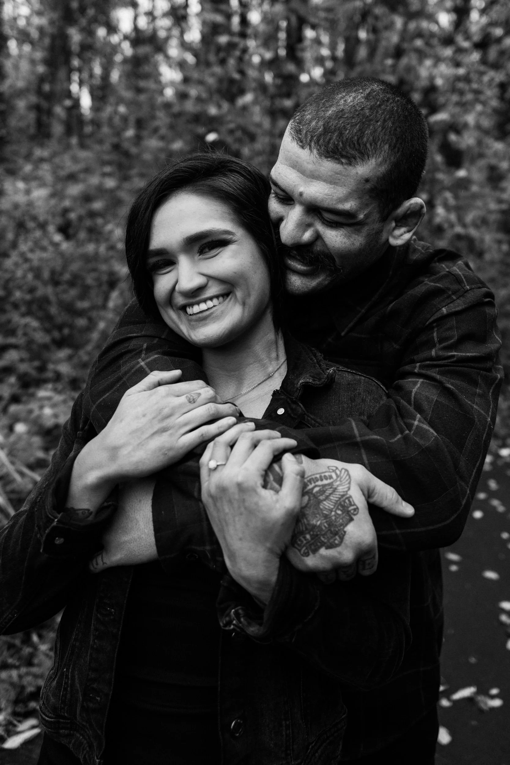 A black-and-white photo of a man and woman smiling and embracing outdoors, with a background of trees and leaves.