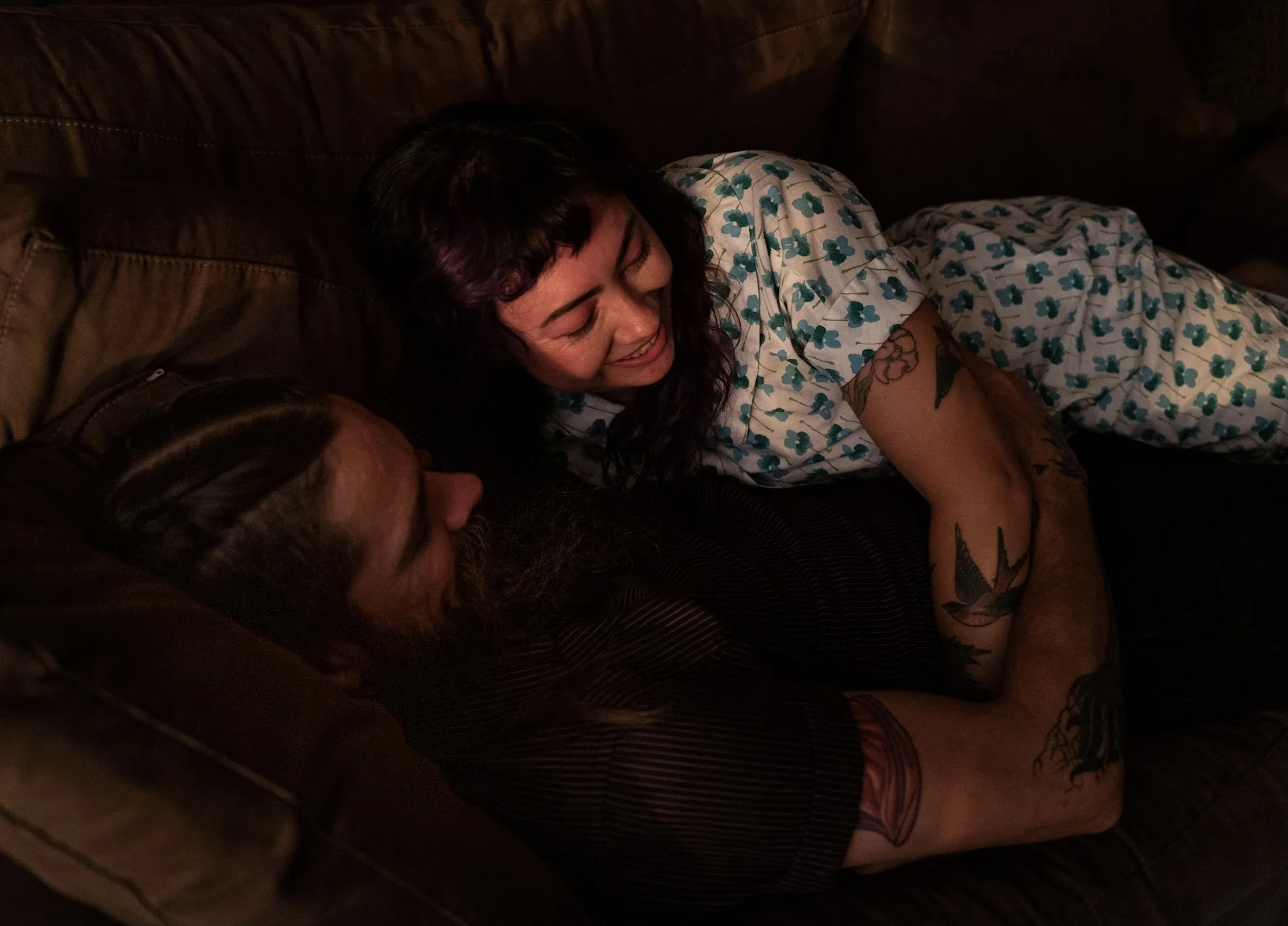 Two people lying on a dark brown couch, smiling and looking at each other. The woman has dark, wavy hair, tattoos on her arm, and is wearing a white shirt with a green floral pattern. The man has a beard and is wearing a dark shirt.