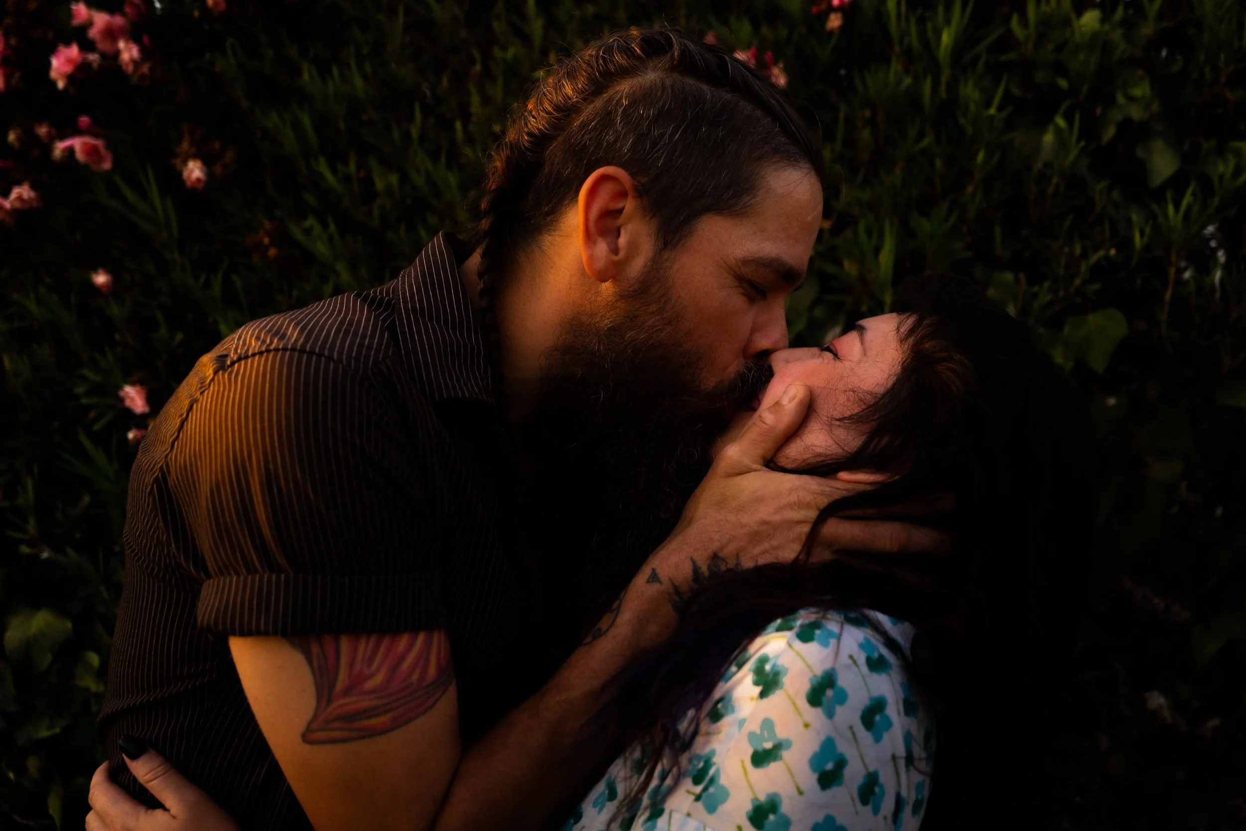 A man with dark hair and a beard is kissing a woman with dark hair and a floral top in front of a background of green plants with pink flowers.