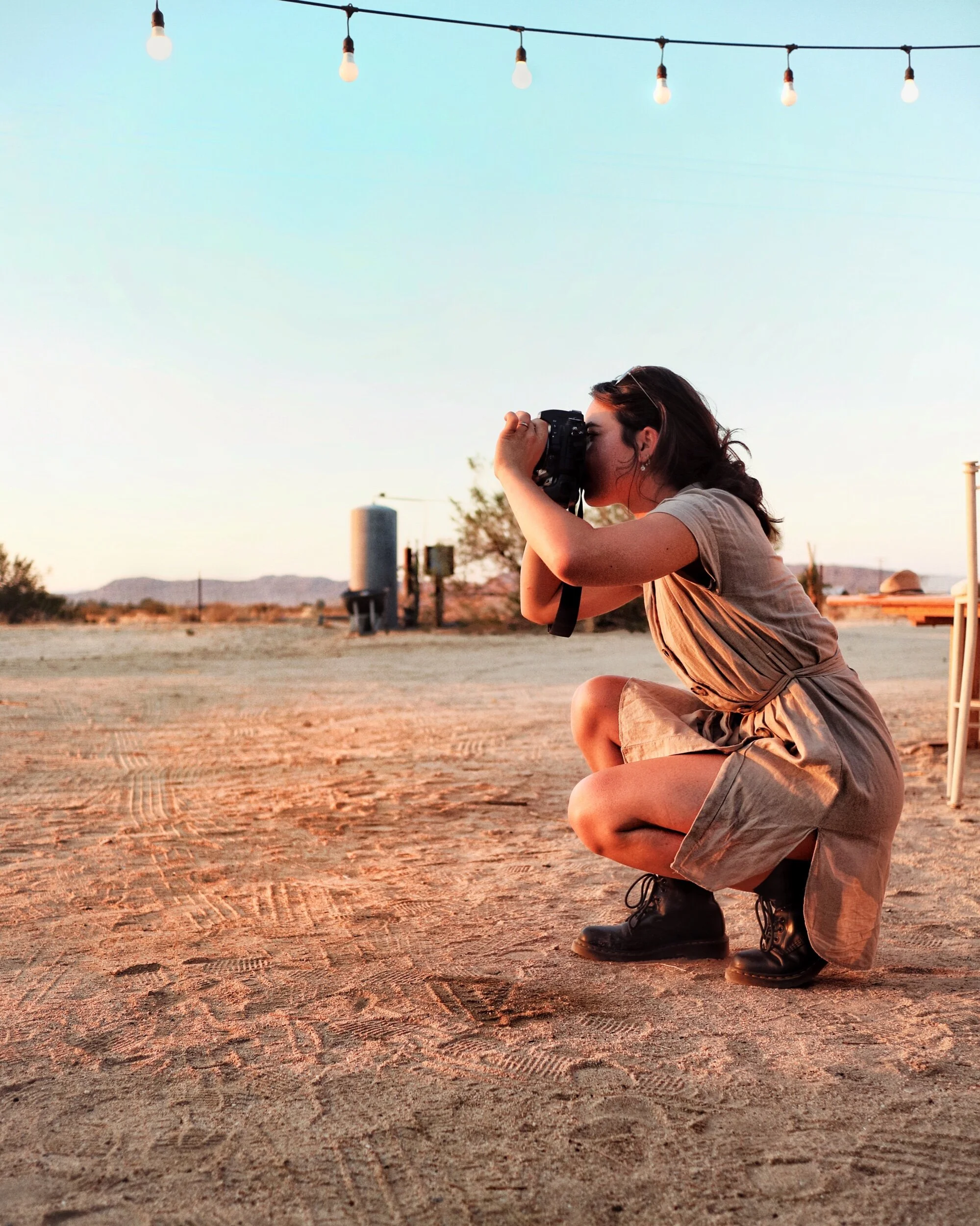 A woman squatting on the ground outdoors, taking a photograph with a camera during sunset. She is wearing a tan dress and black boots, with string lights hanging overhead and a desert landscape in the background.