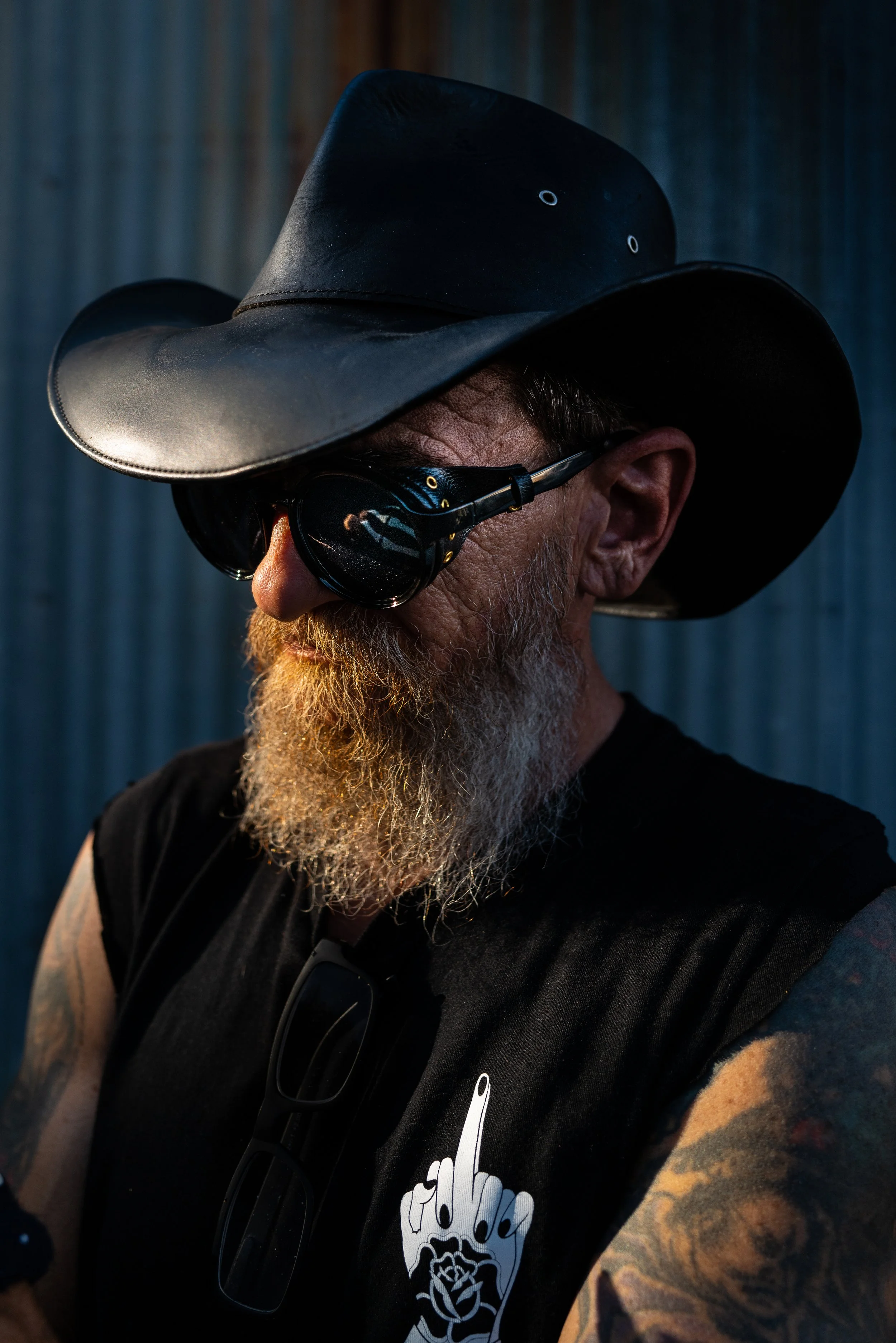 A man wearing a wide-brimmed black hat, sunglasses, and a black sleeveless shirt with a graphic of a hand with an extended middle finger and a rose, showing tattoos on his arm.