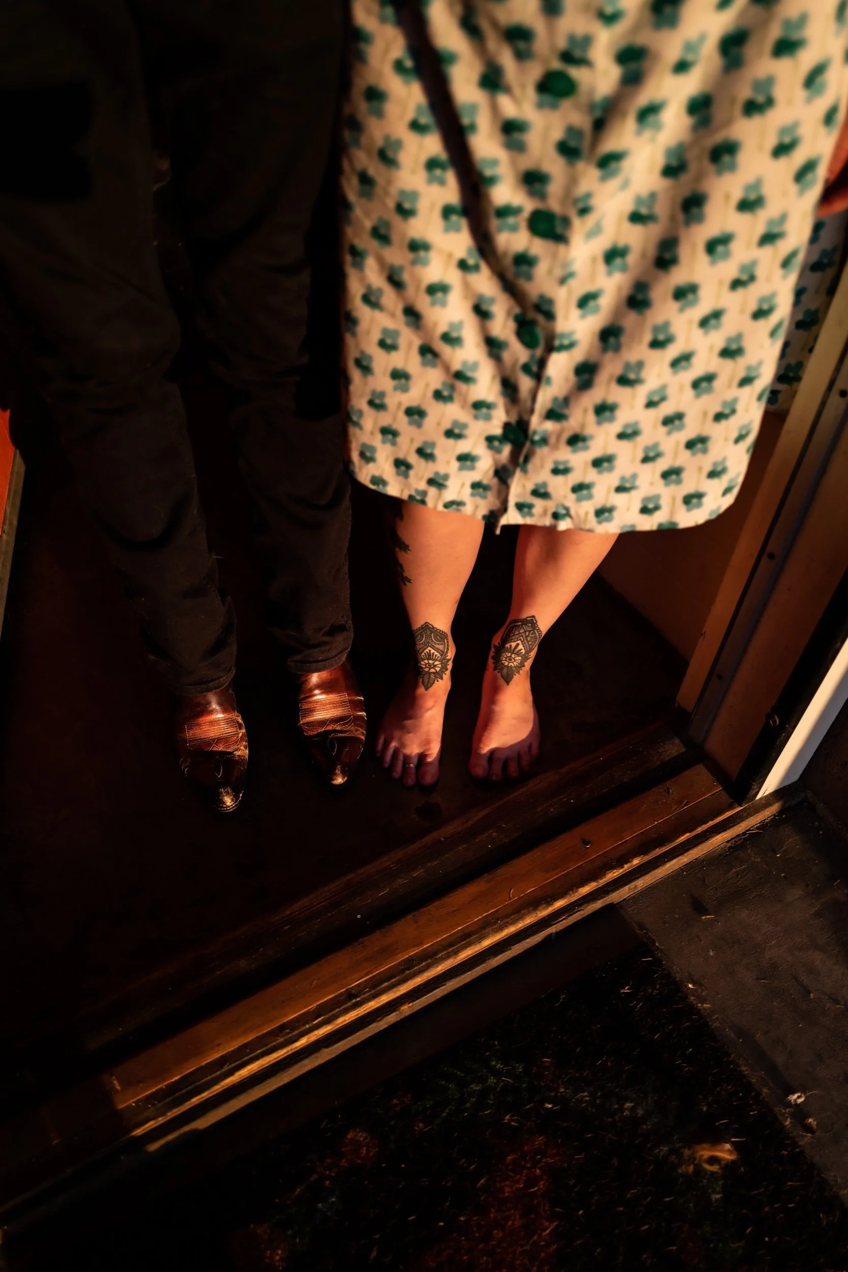 Close-up of two women's legs, one with tattoos, standing on a dark wooden floor in front of a mirror, with a patterned dress and shoes, and another person in black pants and shoes standing beside them.