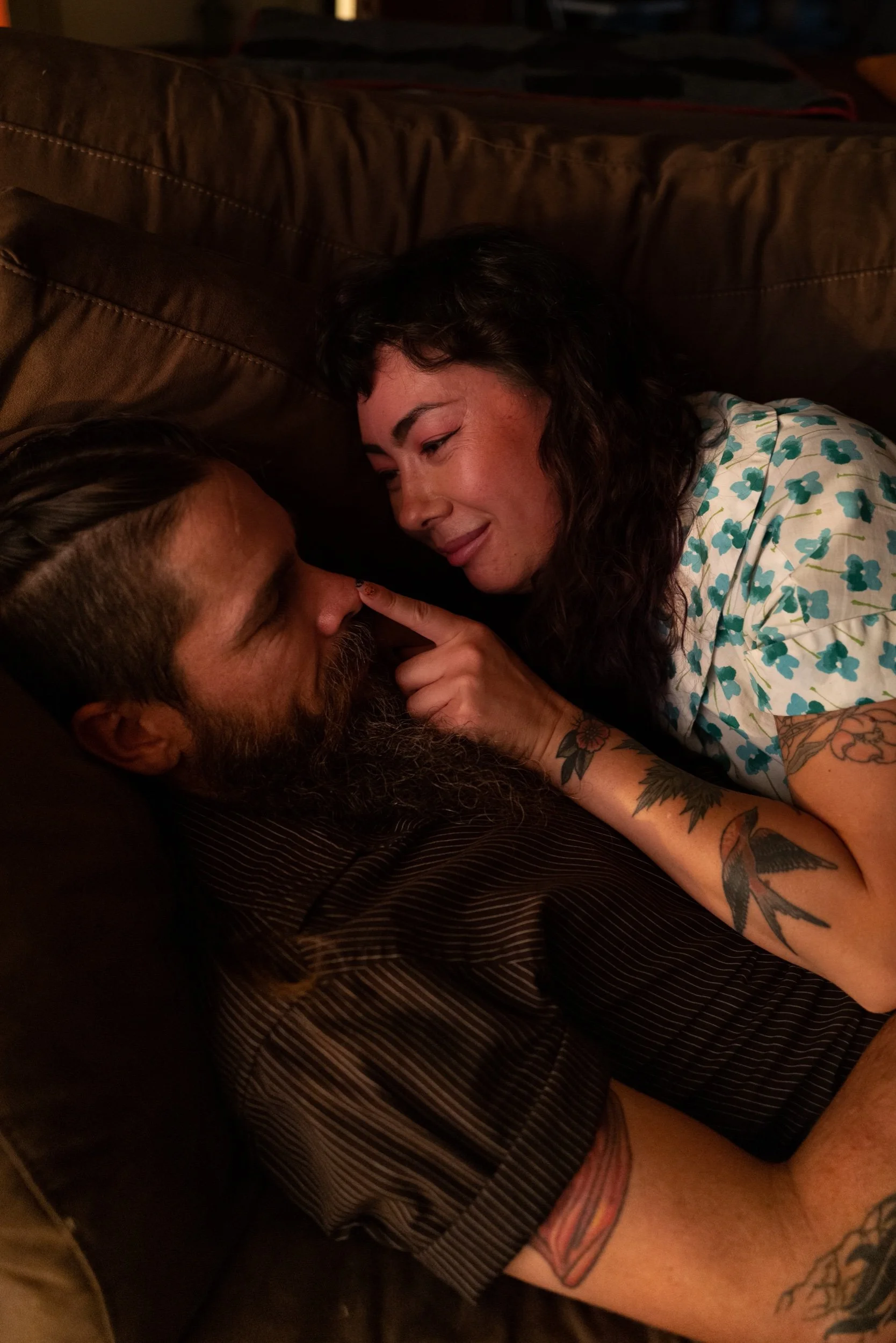 A man and woman lying close together on a brown couch, smiling and touching noses, sharing an intimate moment.