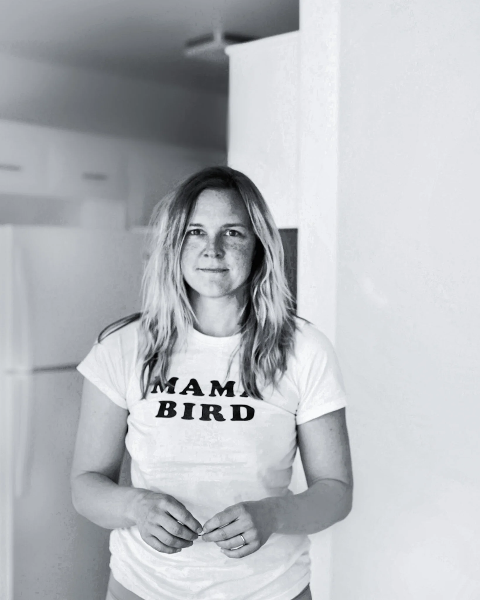 Black and white photo of a woman with shoulder-length hair smiling, wearing a T-shirt that reads 'MAMA BIRD', standing against a plain indoor wall with her hands clasped.