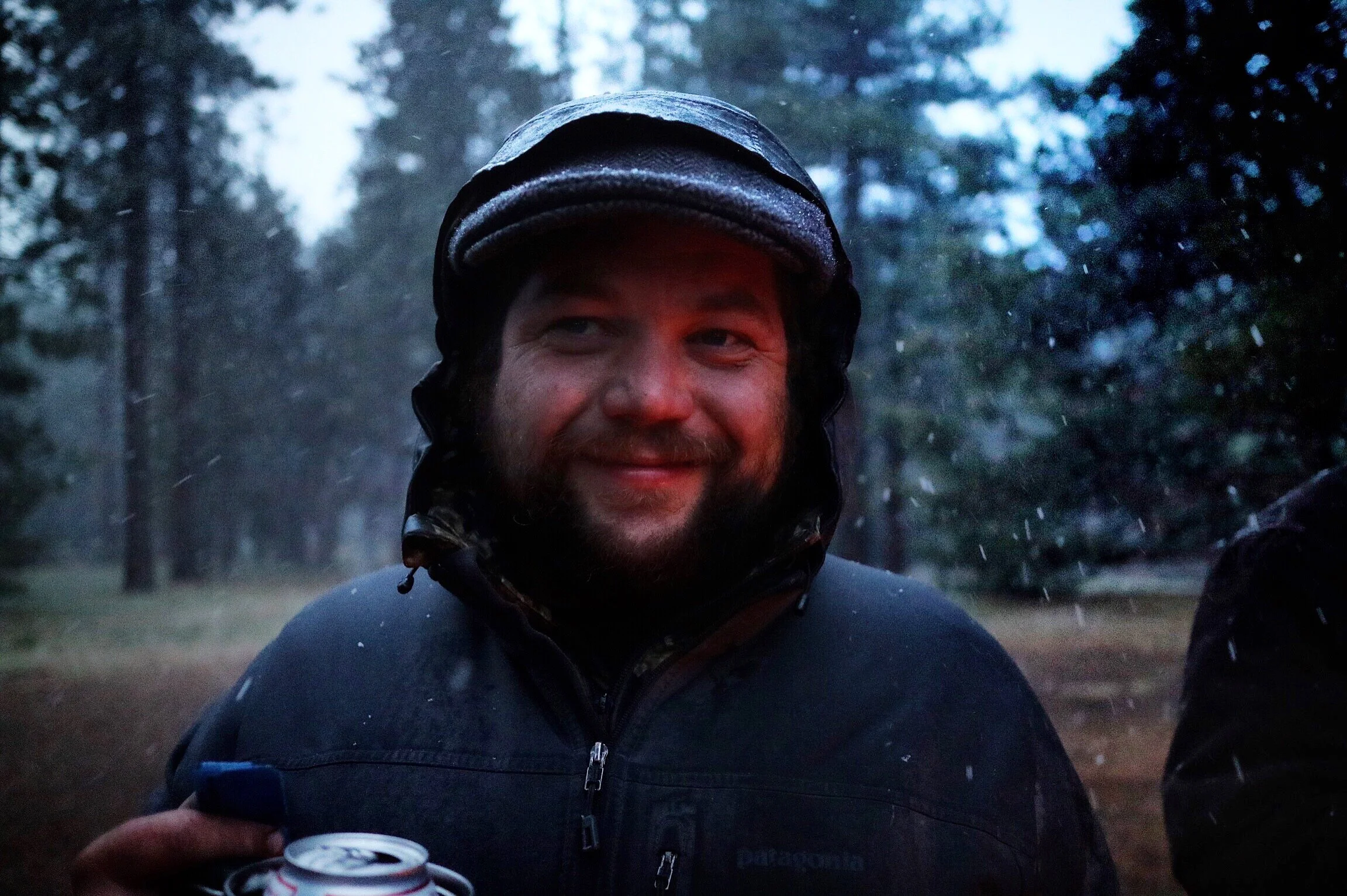 A man wearing a black helmet and a black jacket, smiling, in a forested area during rainy weather, holding a soda can.