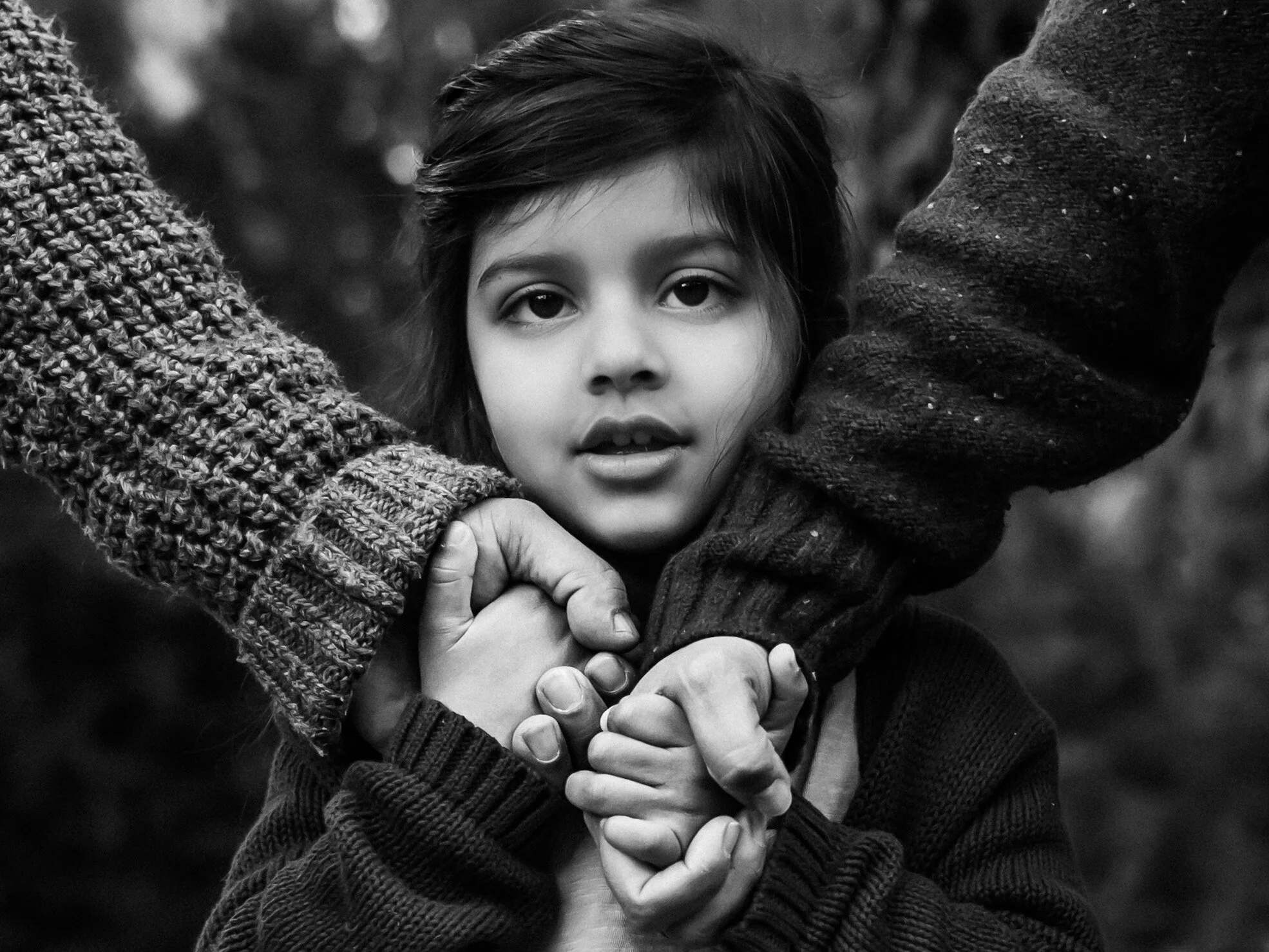 A black and white photo of a young girl with dark hair and expressive eyes, being held and supported by two adults whose hands and arms are visible around her.