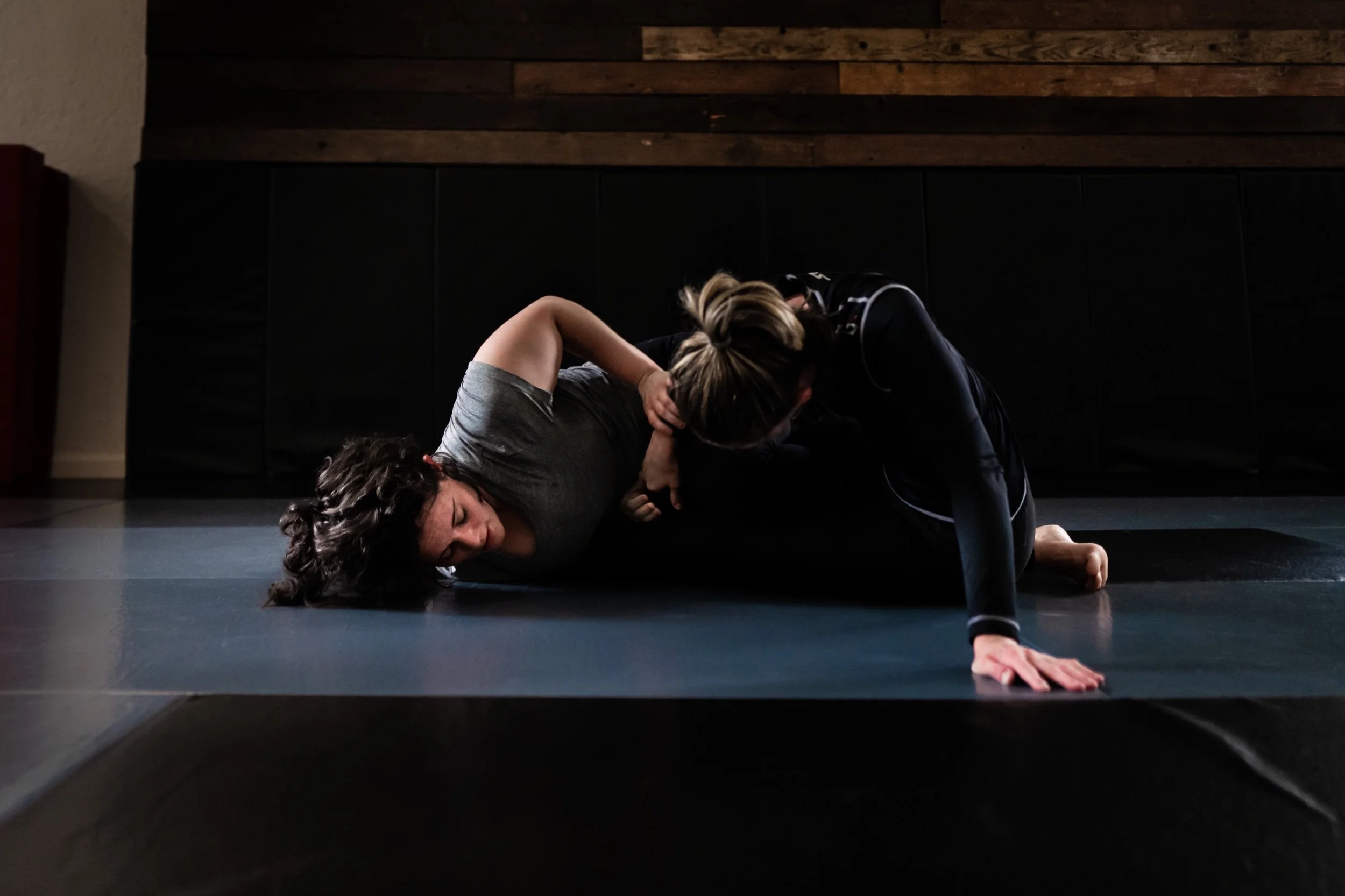 Two women practicing Brazilian Jiu-Jitsu on a black mat in a room with wooden paneling on the wall. One woman is on the ground, and the other is leaning over her in a grappling position.