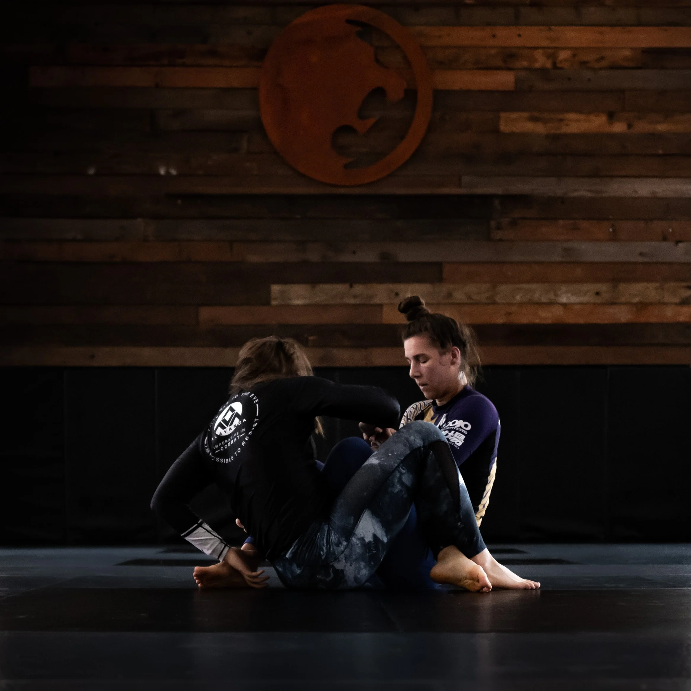 Two people practicing Brazilian Jiu-Jitsu on a black mat in a dimly lit room with wooden wall panels, engaging in a ground fighting technique.