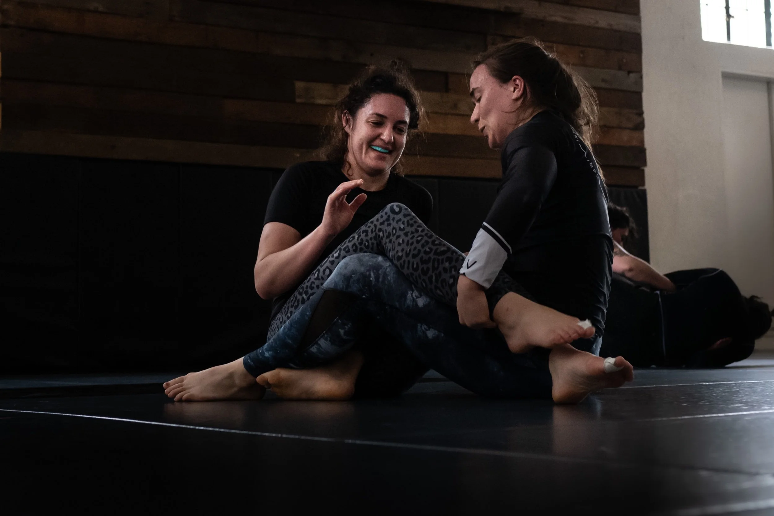 Two women practicing partner yoga on a black mat inside a wooden-finished room, with one woman balancing on the other’s lap and chest, both smiling and engaging in a close, supportive pose.