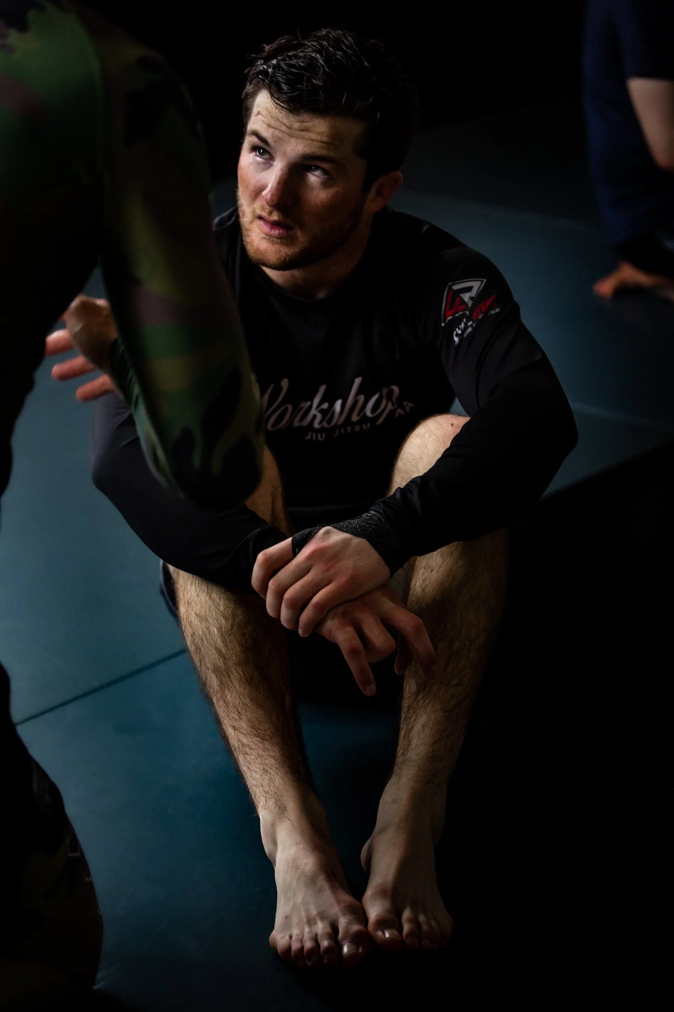 A man with dark hair and a beard, wearing black sportswear with a logo, is sitting on the ground with his legs extended. He is looking upwards at a person in camo clothing. The setting appears to be a martial arts or fitness training environment, wit