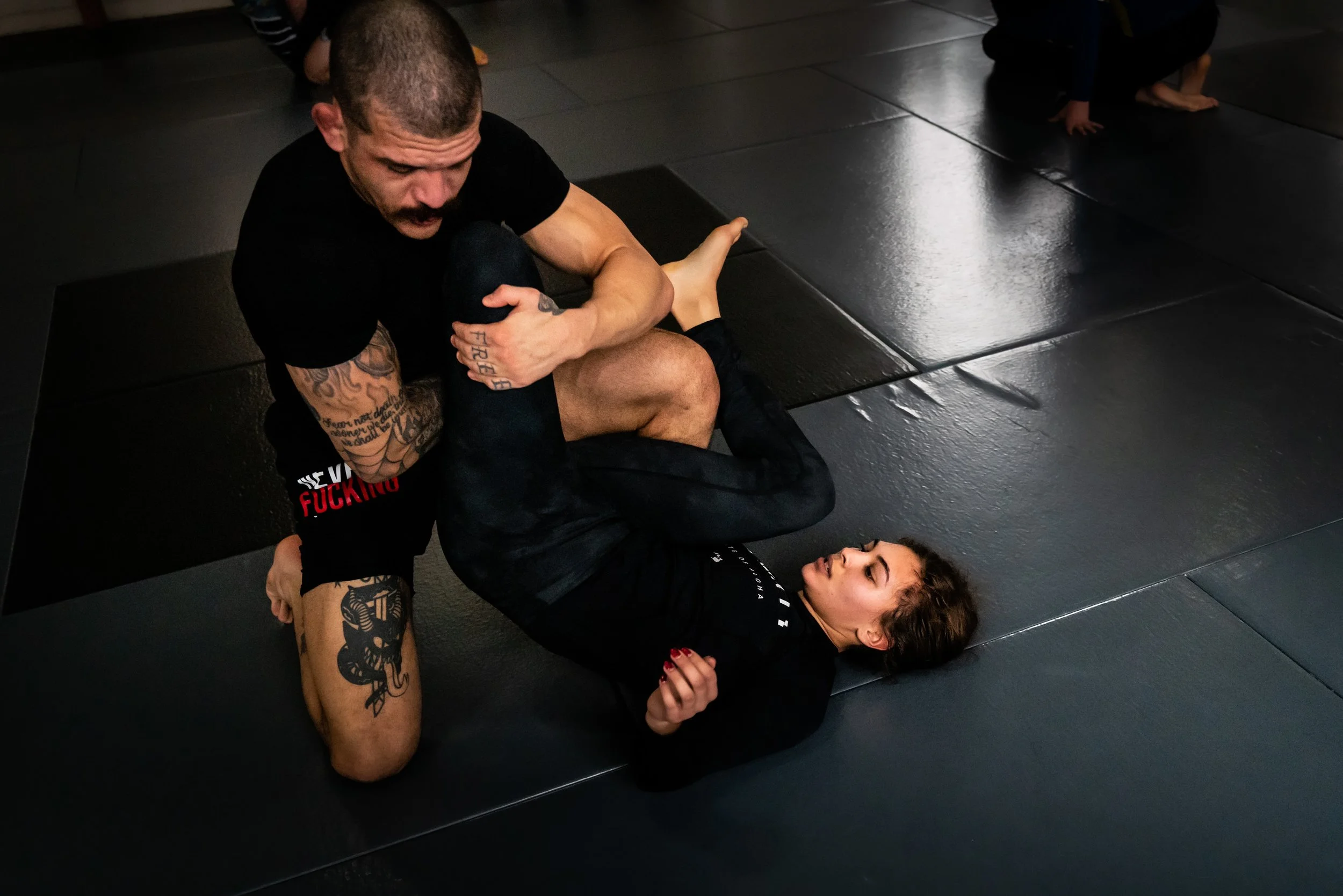 A man with tattoos on his arms and face, wearing a black t-shirt with red and white text, is grappling with a woman on a black mat in a martial arts or Brazilian Jiu-Jitsu class. The woman is on her back, looking up at the man, both engaged in a phys