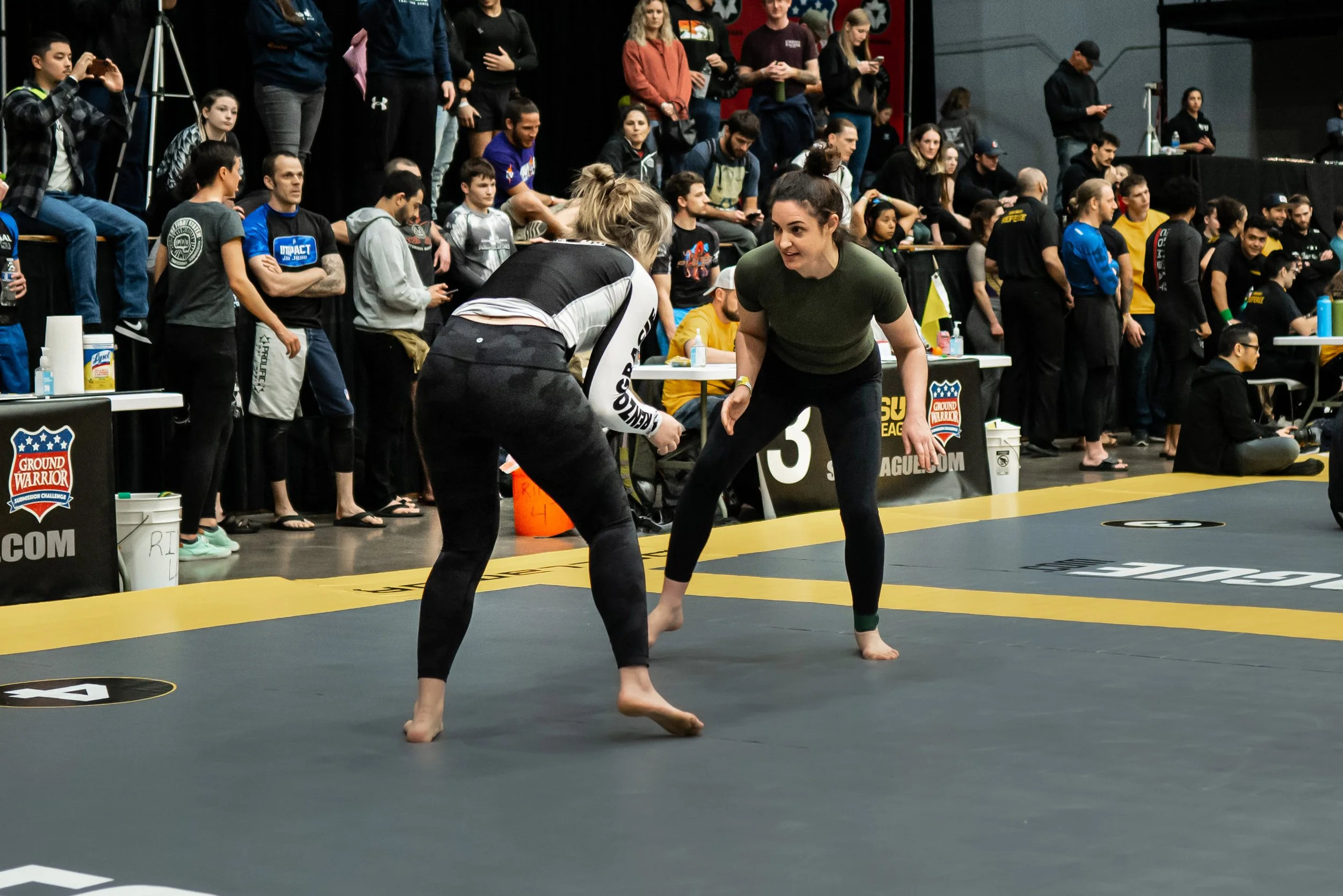 Two women in sportswear competing in a Brazilian Jiu-Jitsu match on a mat with yellow borders, while a crowd of spectators watches from the background.