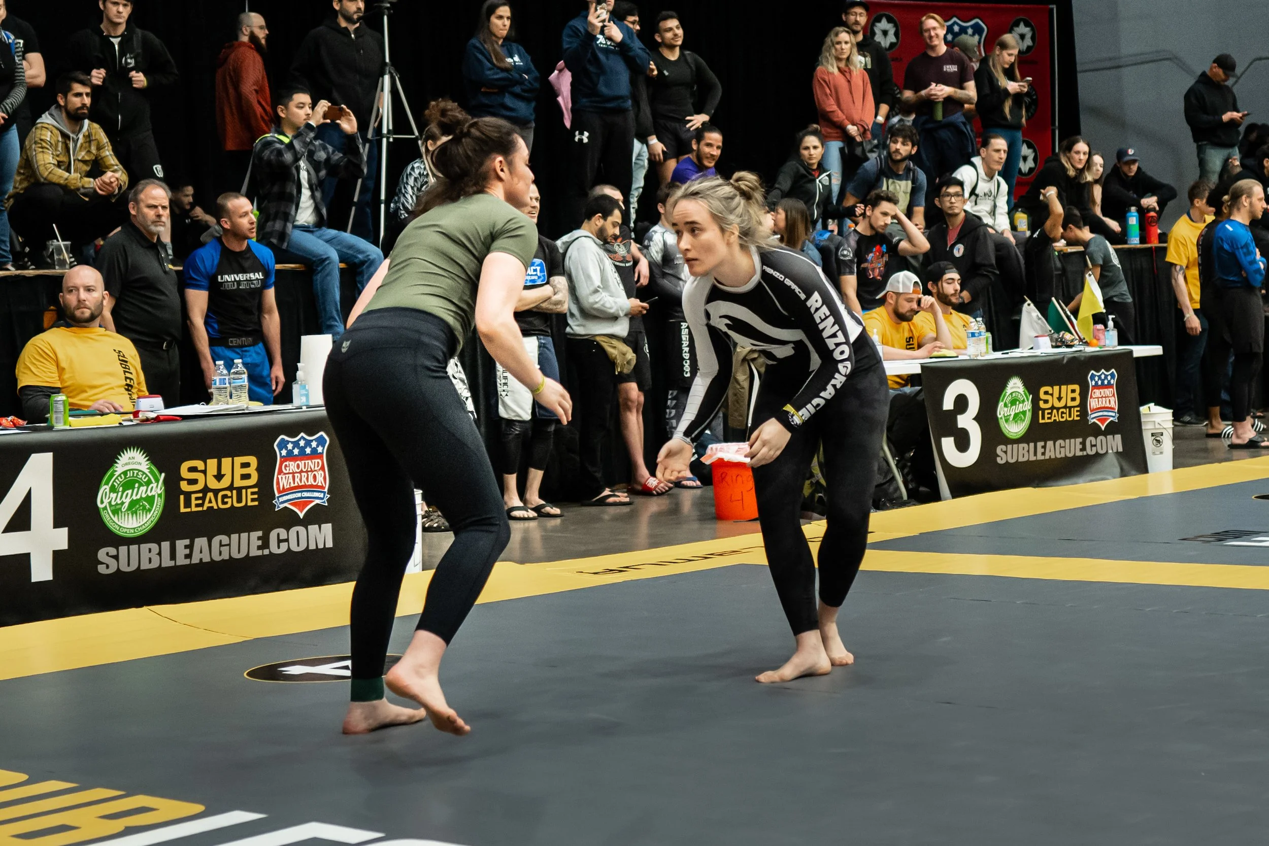 Two women are practicing Brazilian Jiu-Jitsu on a mat at a martial arts competition, with a large audience watching behind a barrier displaying event and sponsor logos.
