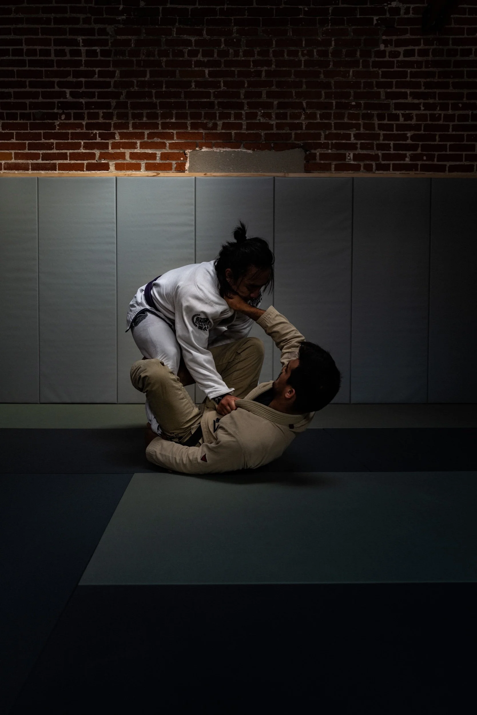 Two individuals engaged in Brazilian Jiu-Jitsu sparring on a mat in a training facility with a brick wall and padded wall panels. One person in a white gi is on top of a person in a beige gi, executing a grappling move.