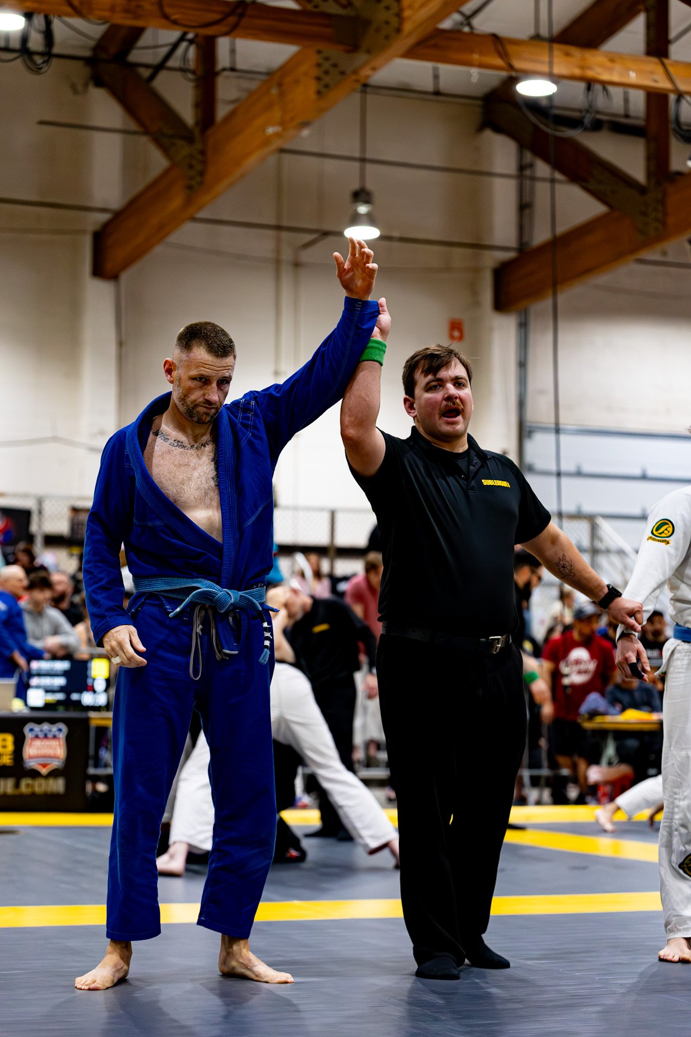 A male Jiu-Jitsu athlete in a blue gi and a male referee in black uniform are holding their arms up after a match in a gymnasium with spectators in the background.