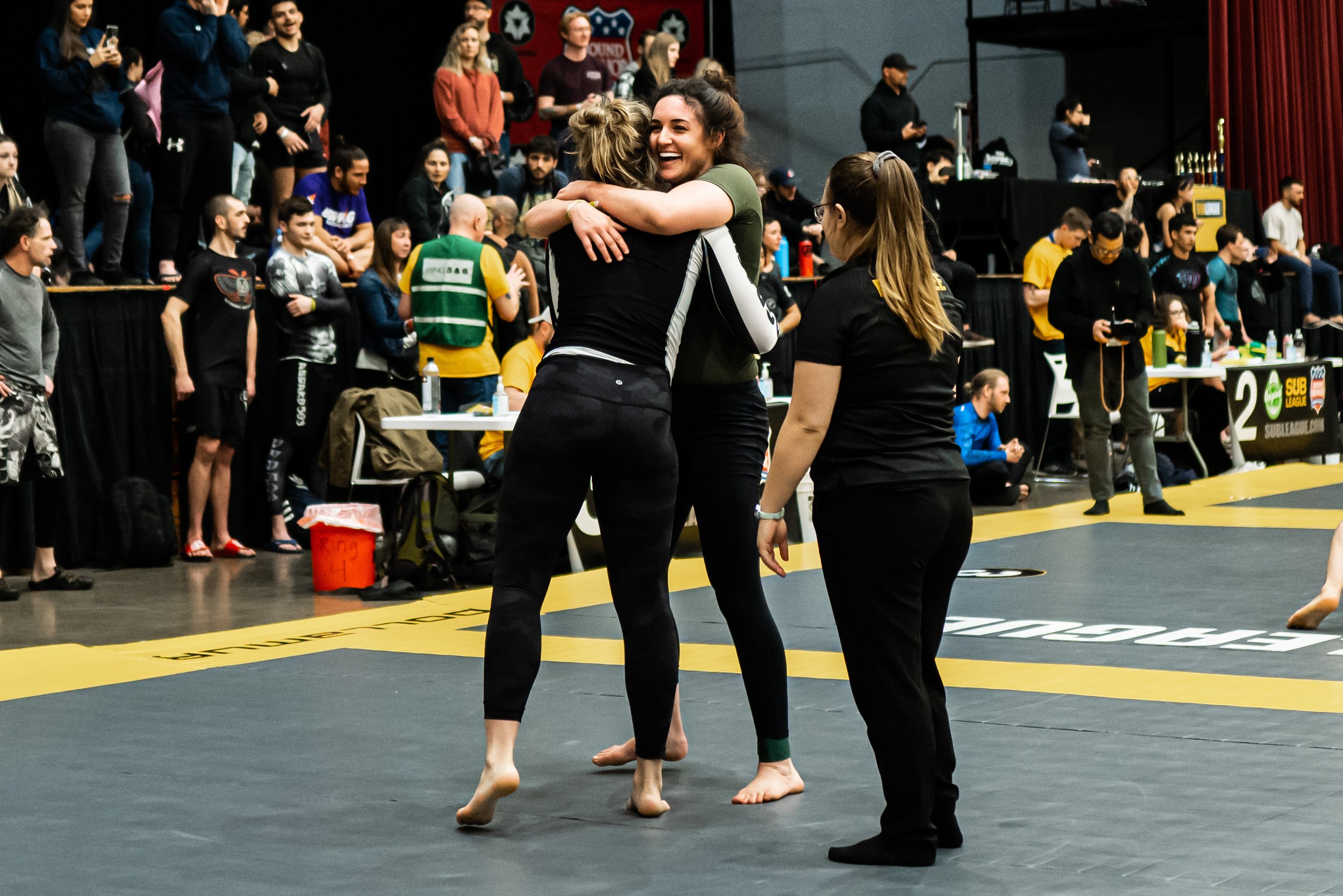 Two women are hugging on a martial arts competition mat, smiling. One woman has curly hair and the other has blonde hair, both in athletic gear. In the background, people are watching, some standing and some seated, with various banners and trophies 