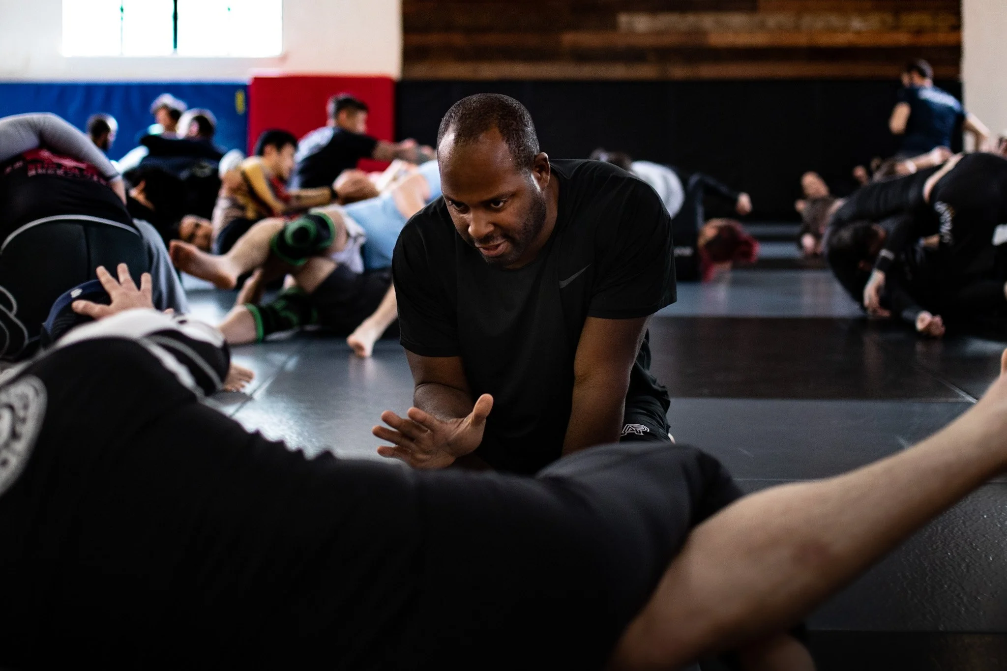 A man in black athletic clothing is kneeling on a gym floor, stretching or practicing a warm-up exercise, with a focused expression. Several other people are in the background, lying or stretching on mats inside a gym or fitness studio, with large wi
