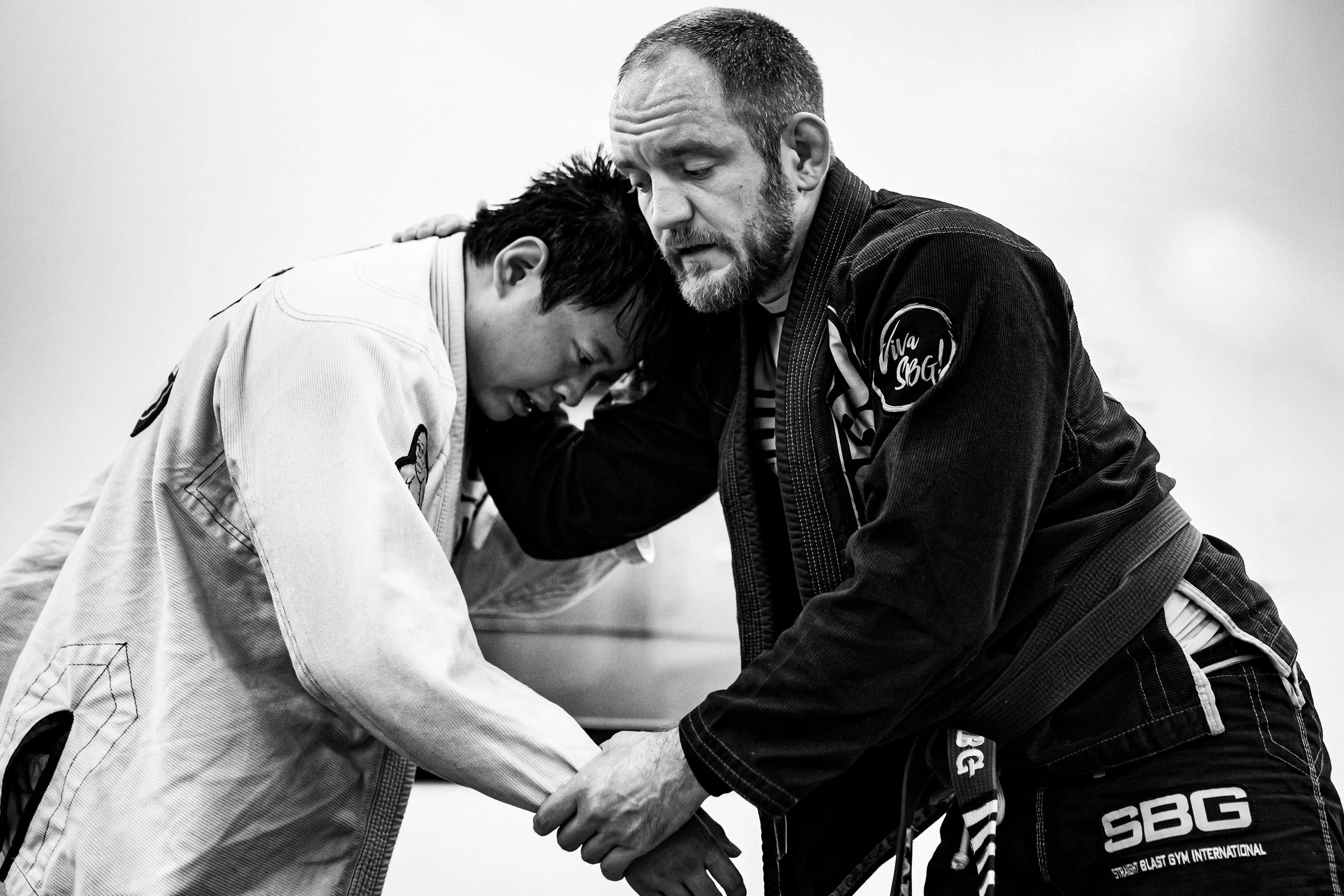 A martial arts instructor demonstrating a technique on a student, both wearing gi uniforms, in a dojo setting.