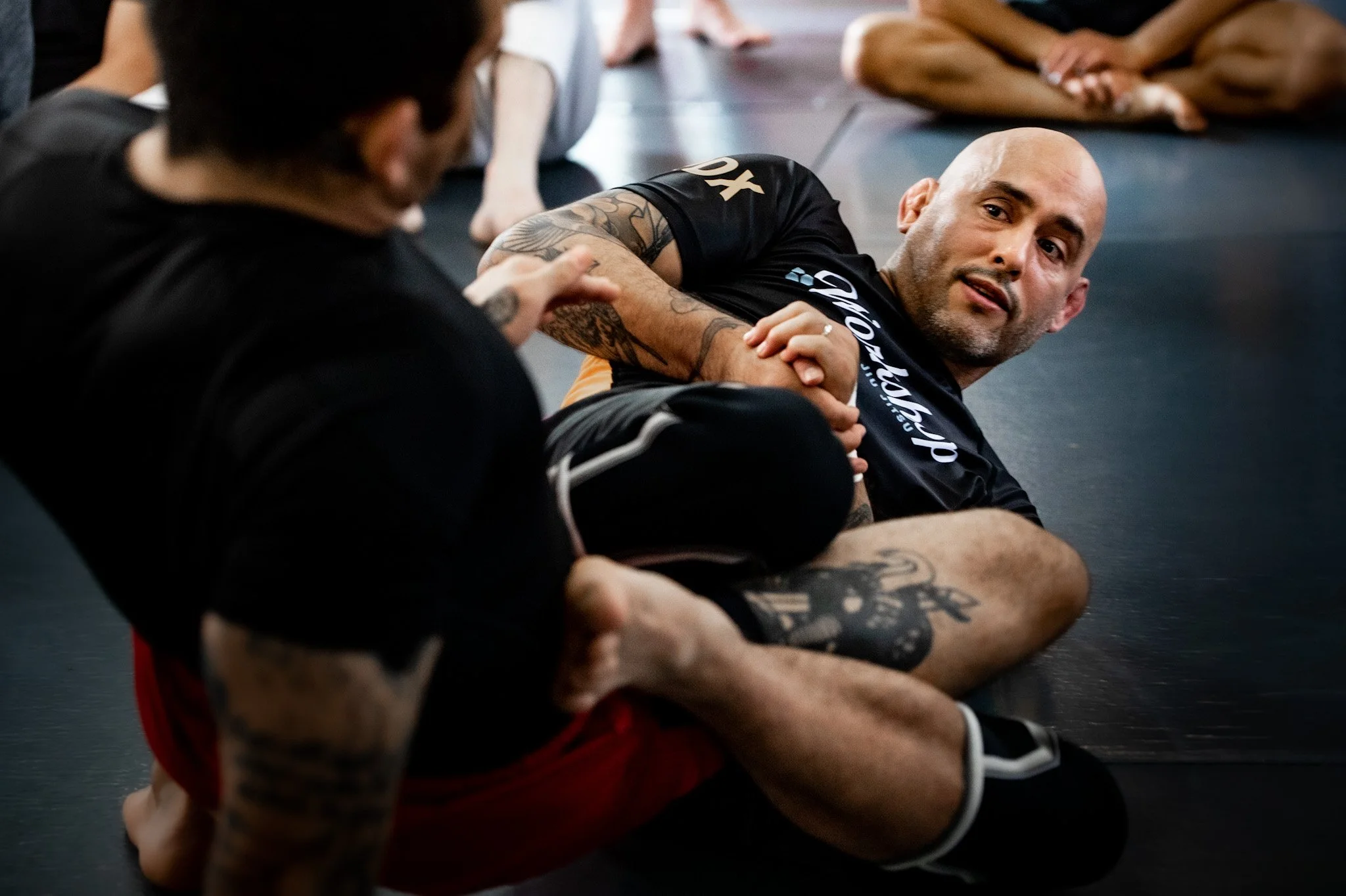 A group of people in athletic clothing participating in a martial arts or self-defense class on the floor. One man with tattoos is looking up, while others are engaged in the activity.