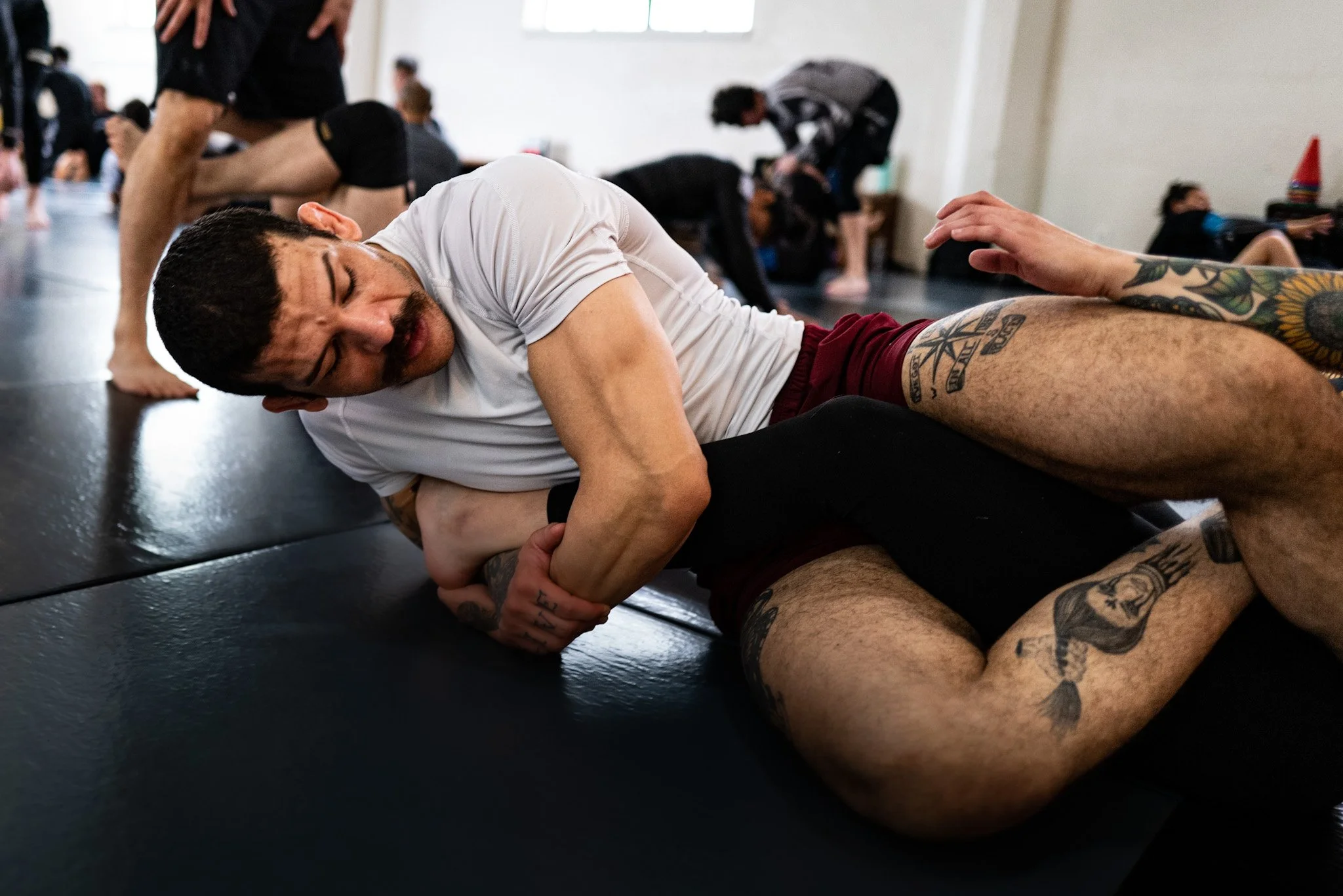 Man with tattoos on his arm and leg practicing Brazilian Jiu-Jitsu or grappling on a black mat, with other participants in the background in a martial arts or grappling class.