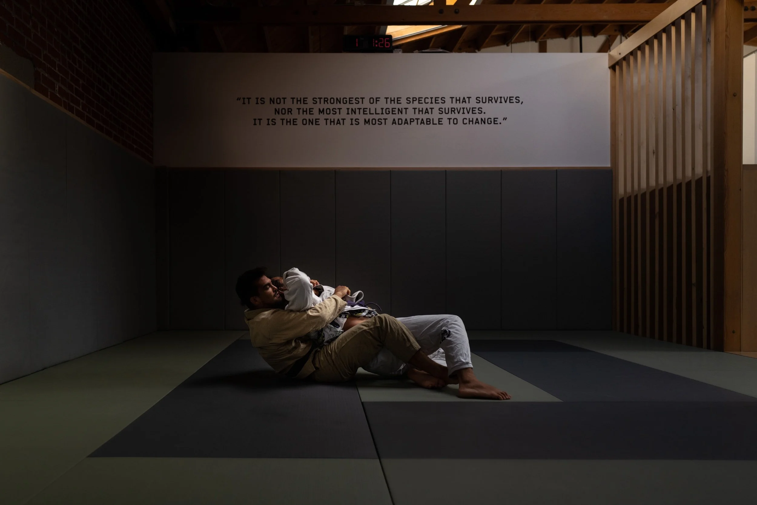 Two brazilian jiu jitsu practitioners in a live round, in the back control position. The setting is a jiu jitsu gym with a sky light and wood beams.