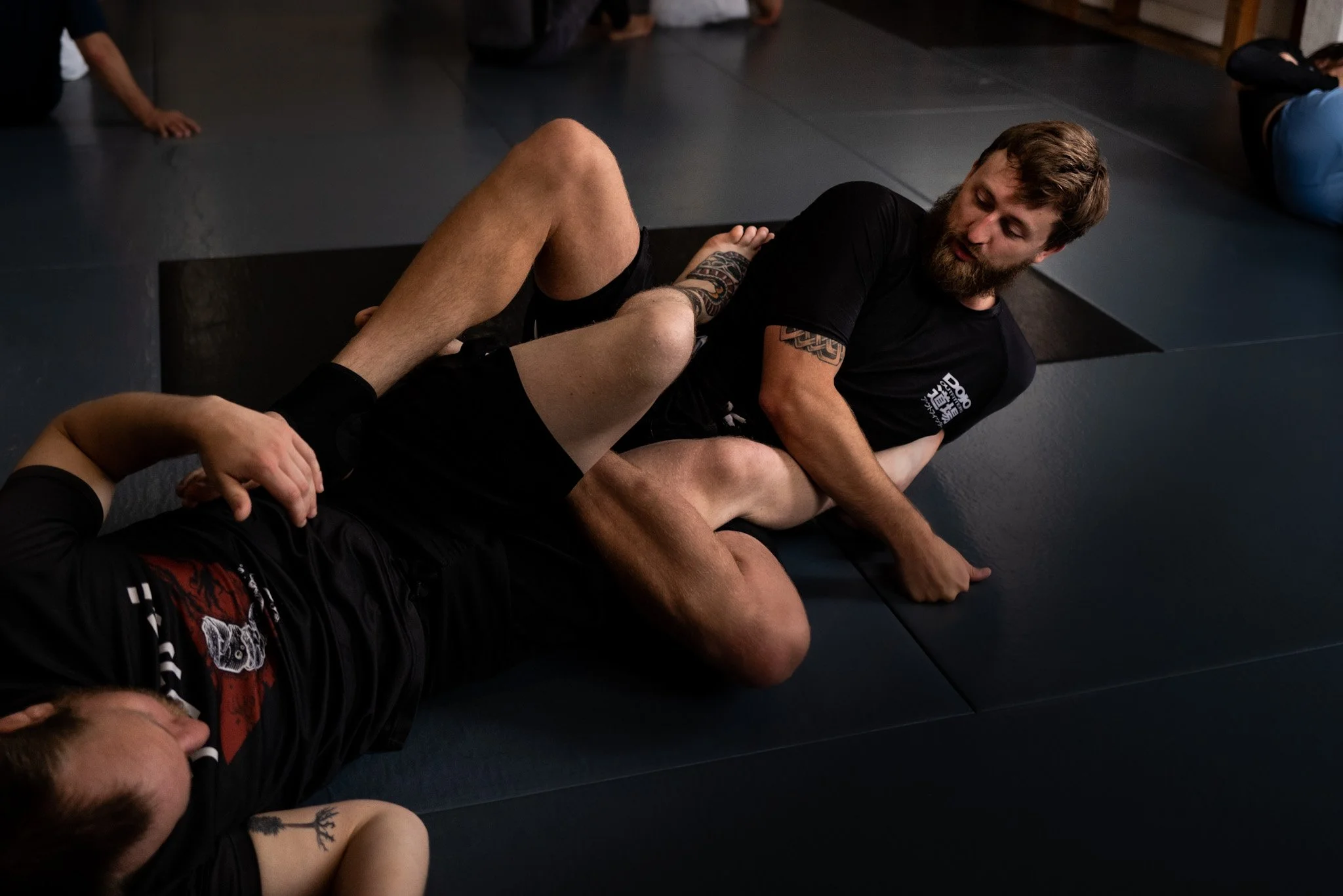 Two men practicing Brazilian Jiu-Jitsu on a black mat, one man is on top of the other, gripping each other's arms and wearing black shirts and shorts.