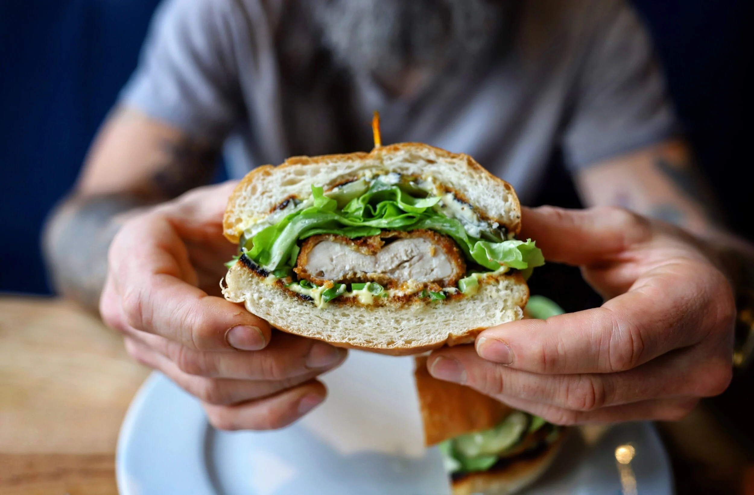 Close-up of a person holding a sandwich with fried chicken, lettuce, and sauce against a blurred background.