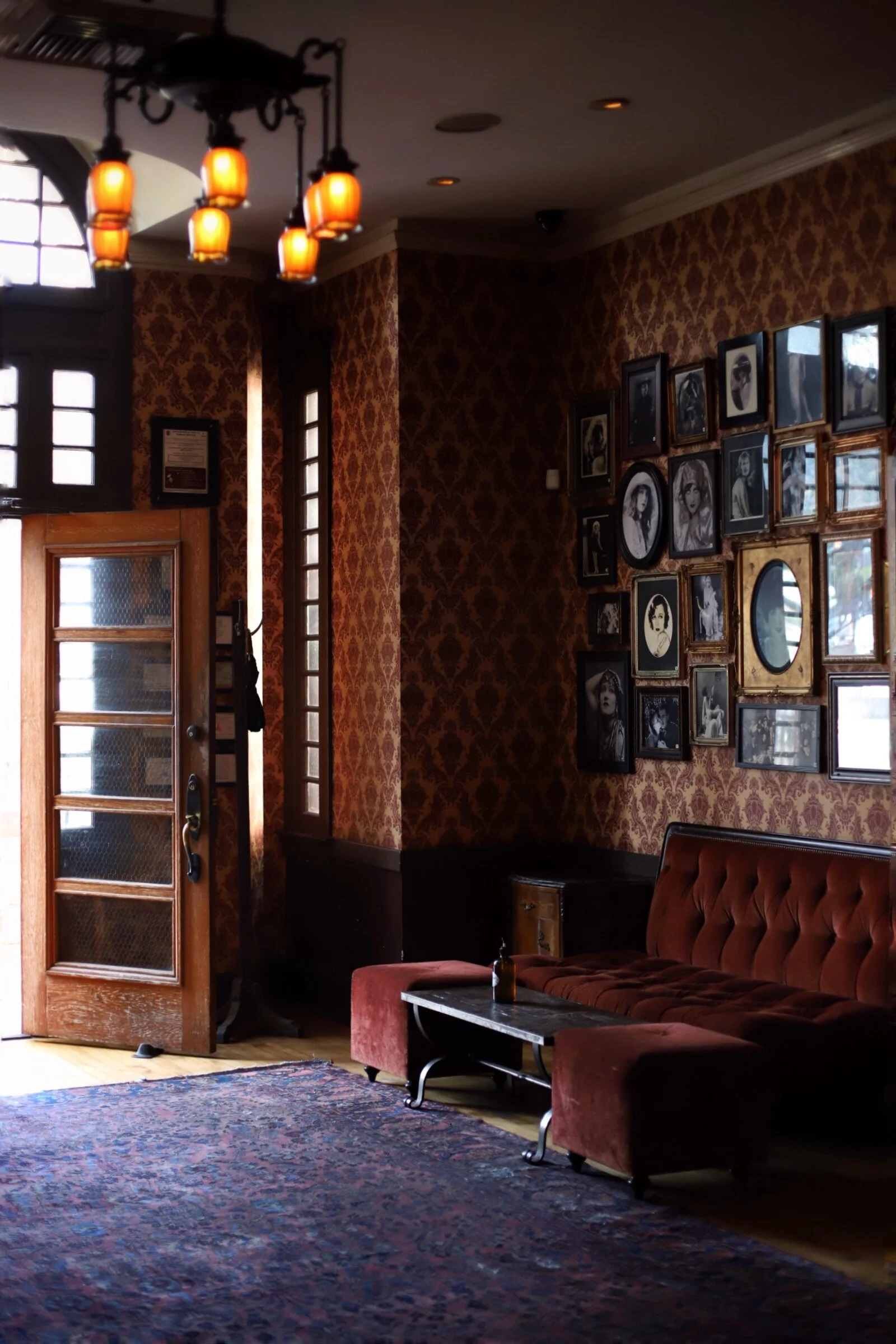Vintage-style room with a red velvet sofa, patterned wallpaper, framed black and white photos, wooden door, and warm lighting.
