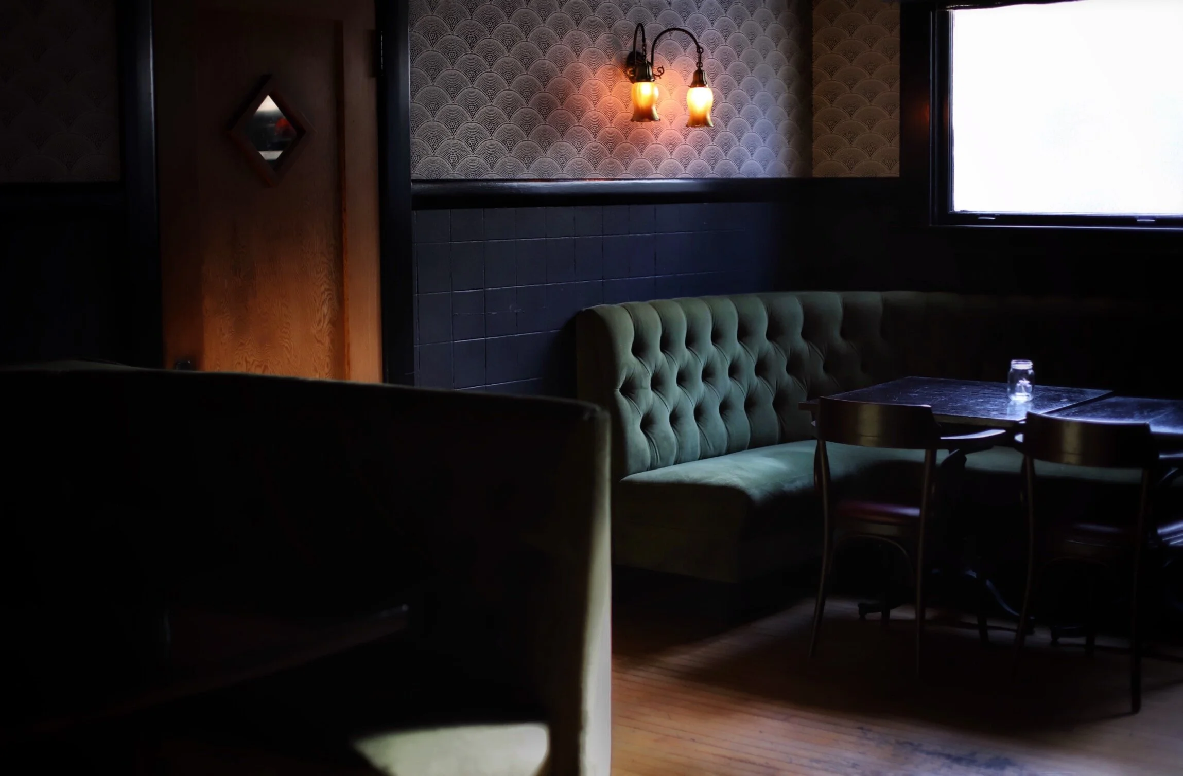 Dimly lit restaurant interior with a green tufted bench, tables, wall-mounted lamps, patterned wallpaper, and a large window.