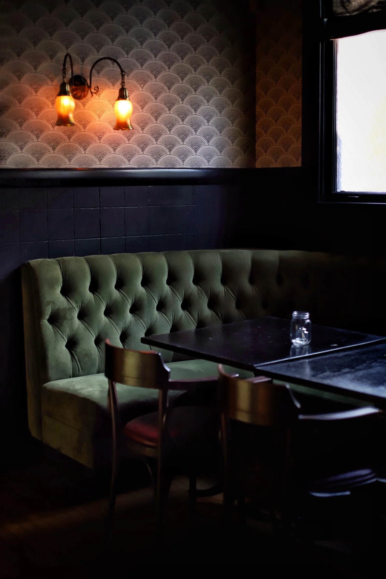 Dimly lit interior of a cafe or restaurant with a green upholstered booth, black marble table, wooden chair, patterned wallpaper, and a window letting in natural light.