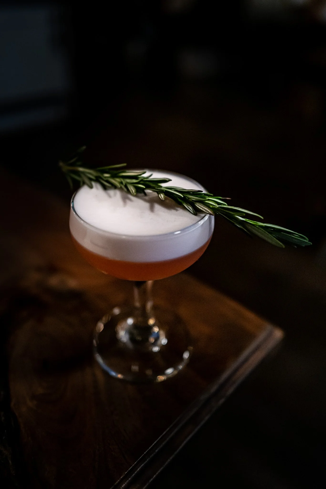 A cocktail in a coupe glass with a sprig of fresh rosemary on top, placed on a dark wooden surface.
