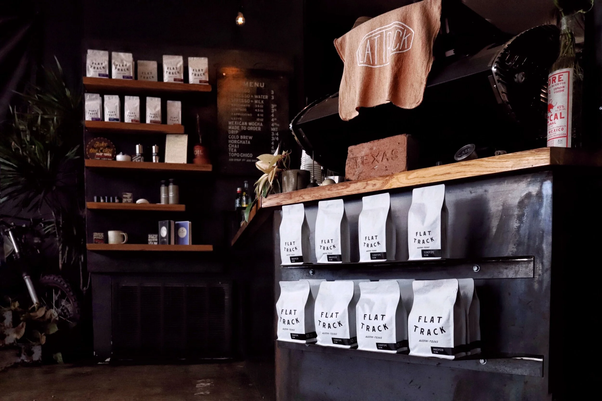 Interior of a coffee shop with bags of coffee labeled 'Flat Track' hanging underneath a counter, a menu on a chalkboard, shelves with coffee products, and a curtain with a napkin bearing the word 'Attack' embroidered on it.