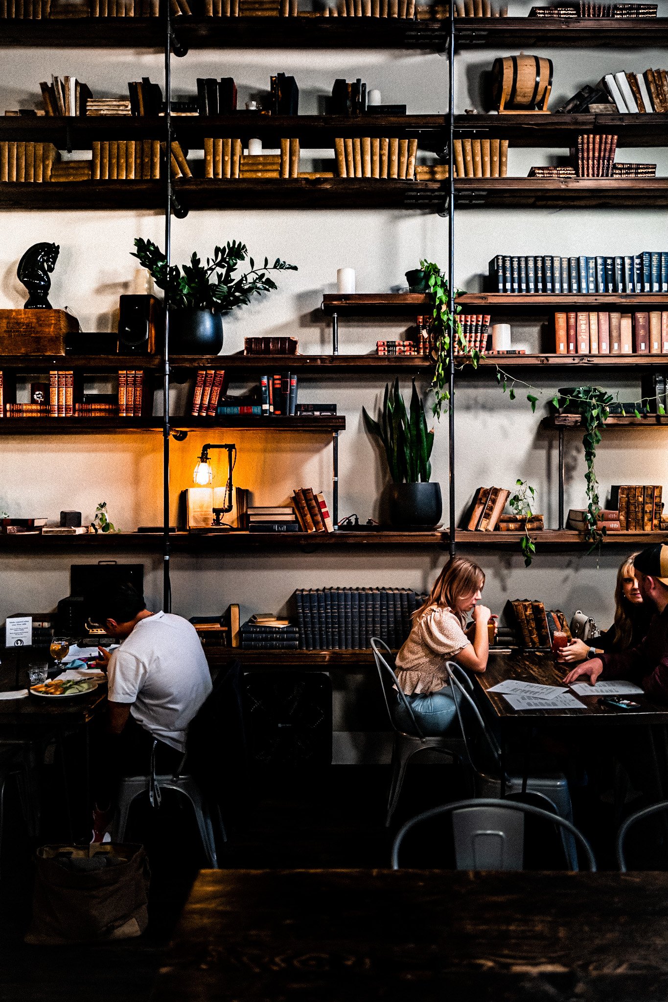 People sitting at tables in a cozy, bookshelf-lined restaurant or cafe, with plants and warm lighting.
