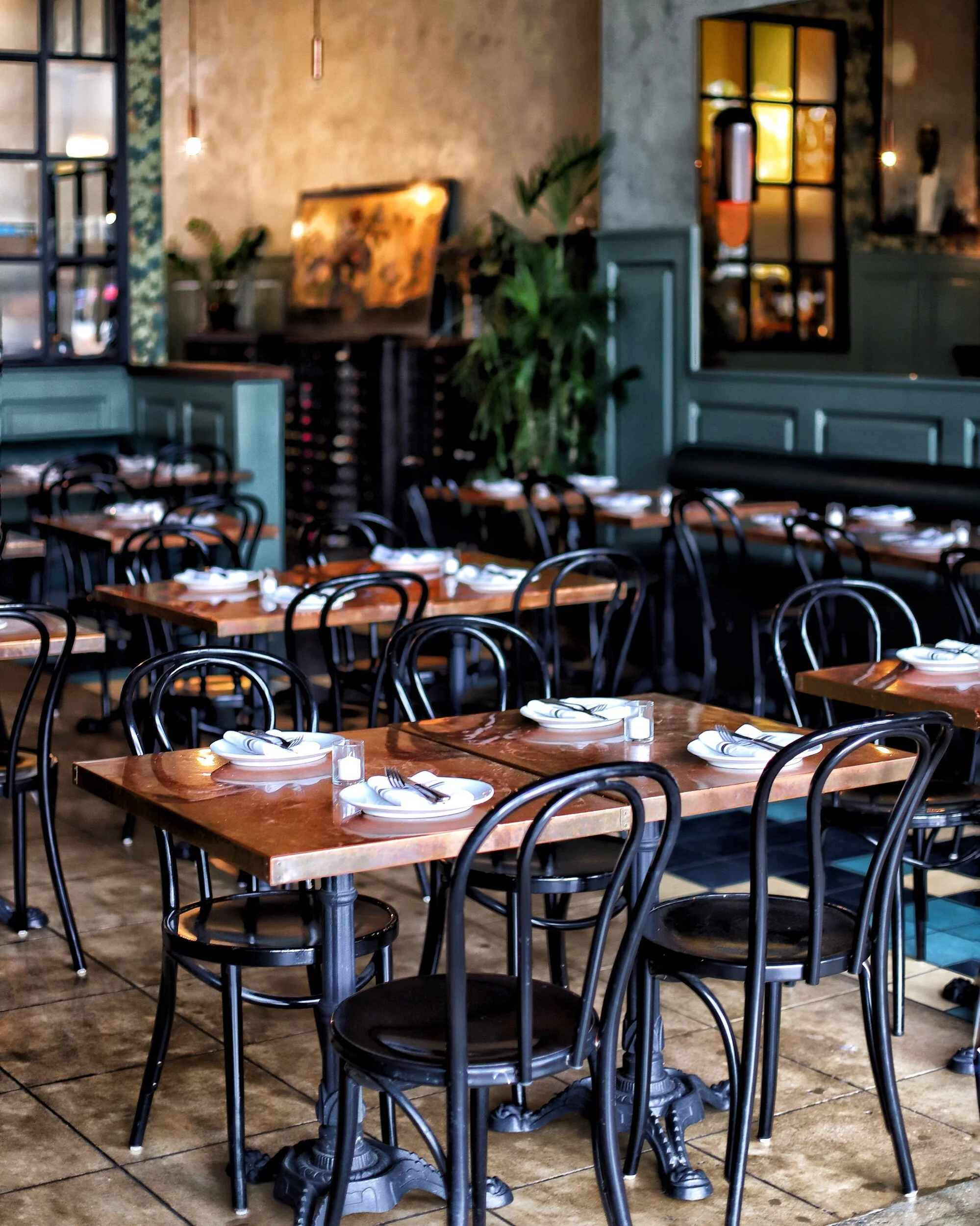 A cozy restaurant interior with wooden tables set with plates and napkins, black chairs, plants, textured walls, and large windows letting in natural light.