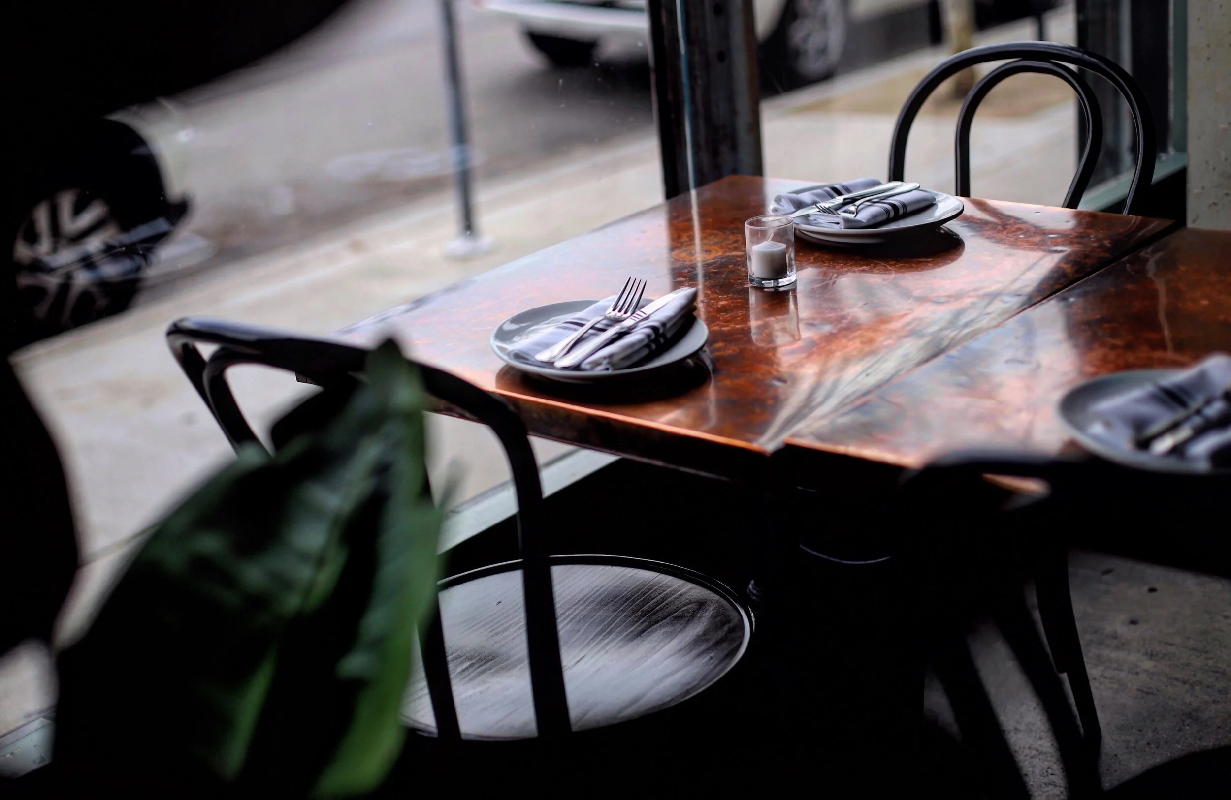 A table set for two with plates, napkins, silverware, a candle, and glasses inside a restaurant with a view of the sidewalk outside.
