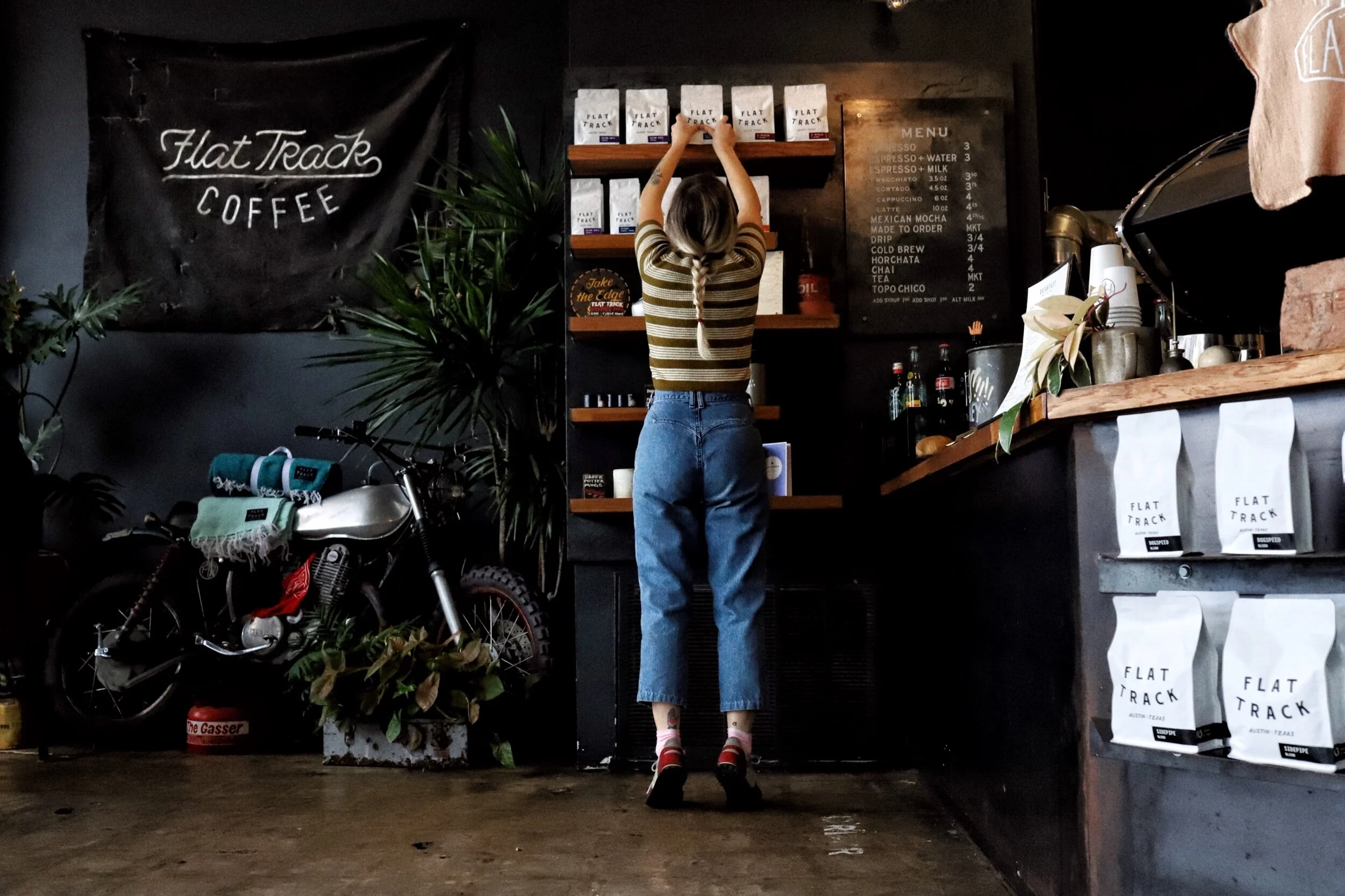 A woman with long hair tied in a braid, wearing a striped t-shirt, jeans, and sandals, is reaching up to grab a bag of coffee from a shelf in a coffee shop. The shop has a black wall with a menu and branded coffee bags, a large green plant, and a vin
