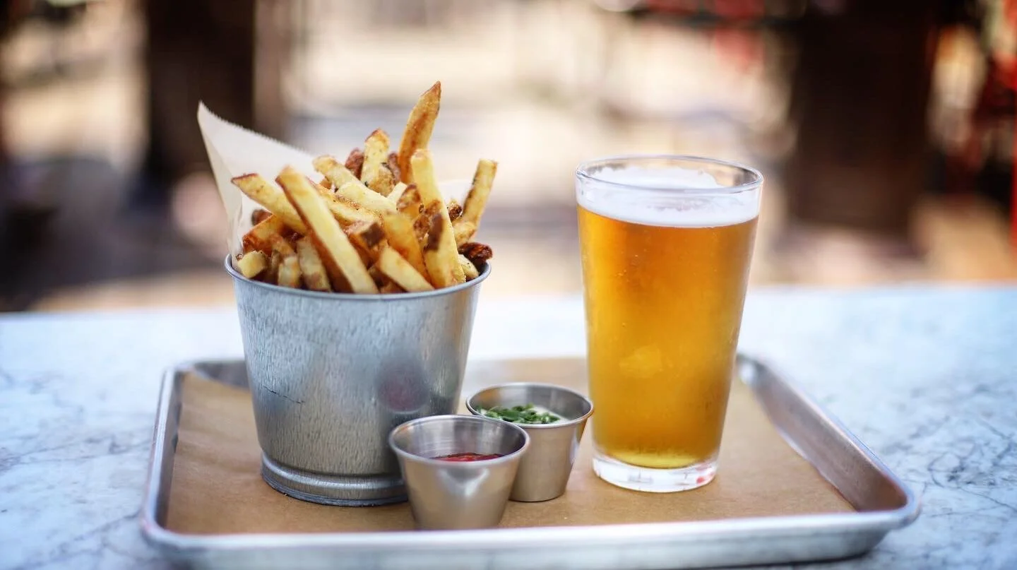 A metal bucket of French fries with three small containers of ketchup, green sauce, and dark sauce, a tall glass of beer, all on a tray atop a marble table.