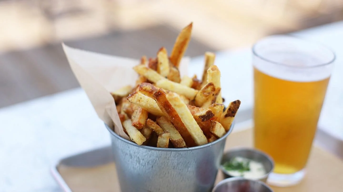A metal bucket filled with French fries on a table, with a glass of beer and small containers of dipping sauces in the background.