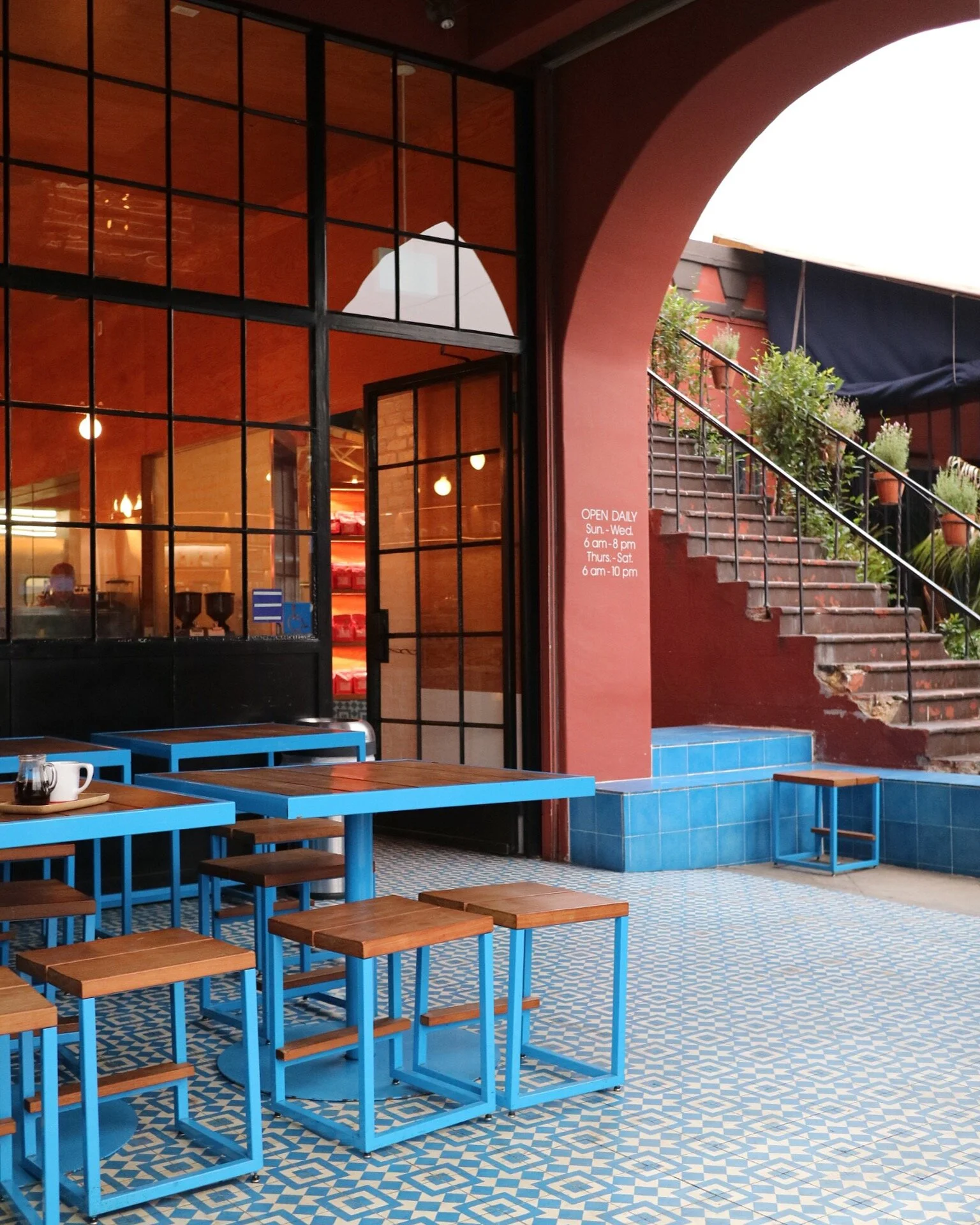 Interior of a restaurant with blue tables and stools, a mosaic tile floor, a brick staircase with plants, and large windows.