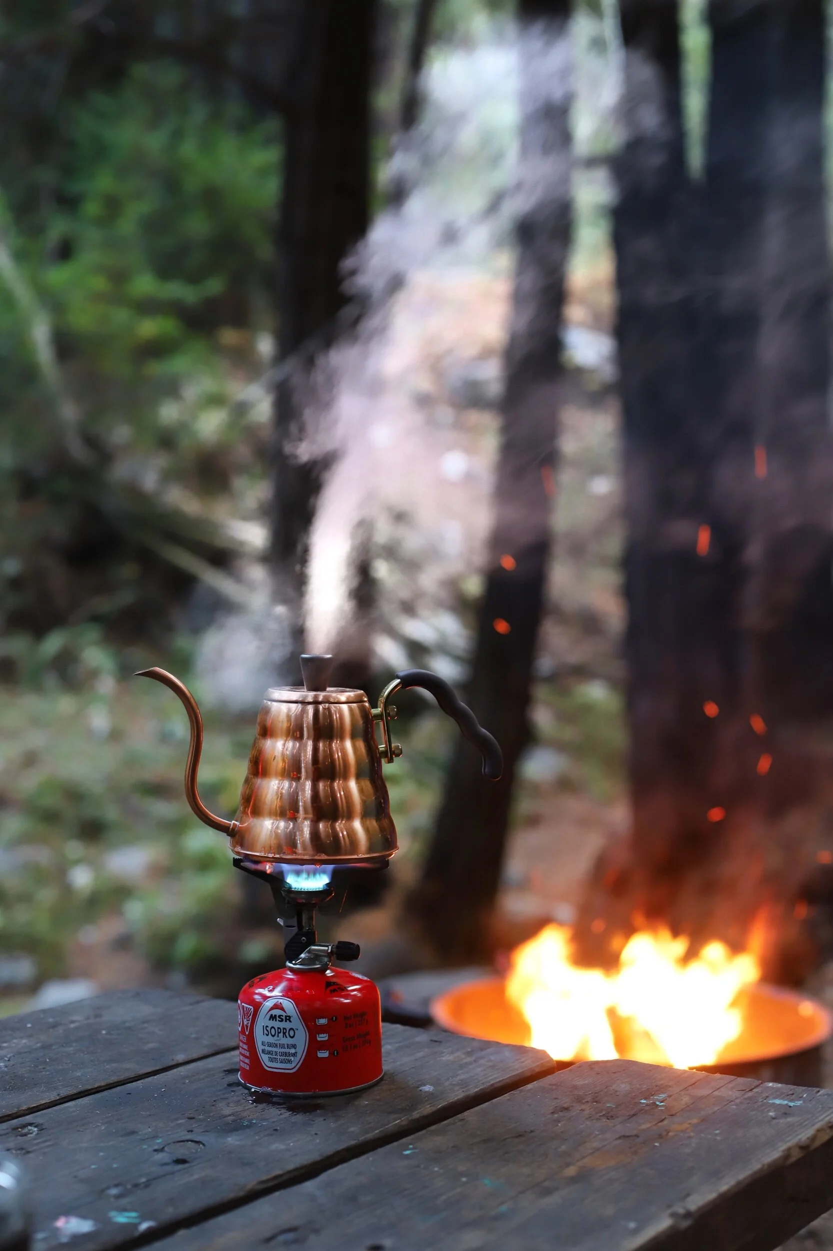 A copper-colored gooseneck kettle on a portable stove, with a flame underneath, set against a blurred outdoor background with trees and smoke.