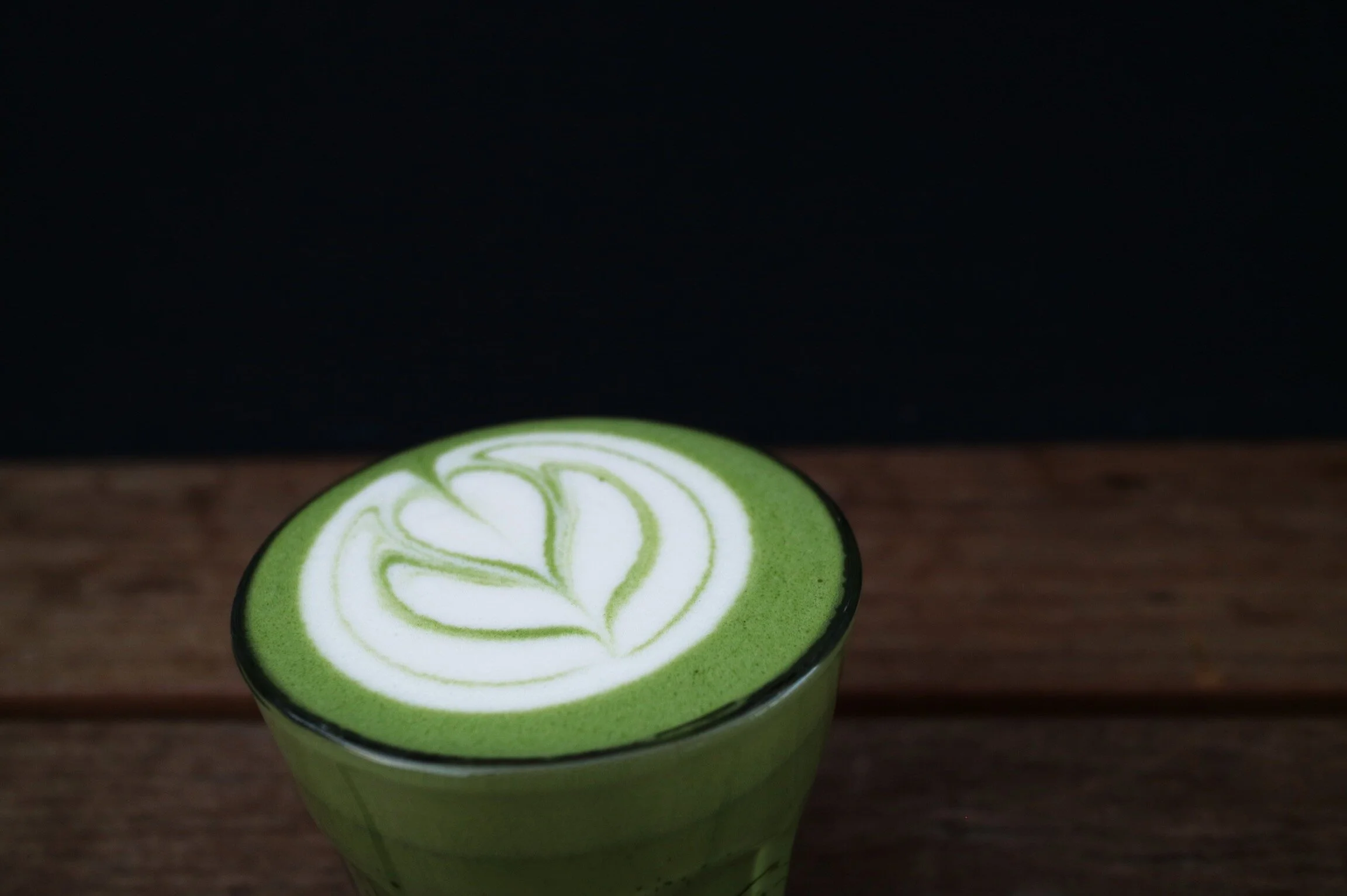 Close-up of a green matcha latte with latte art in a clear glass, placed on a wooden surface.
