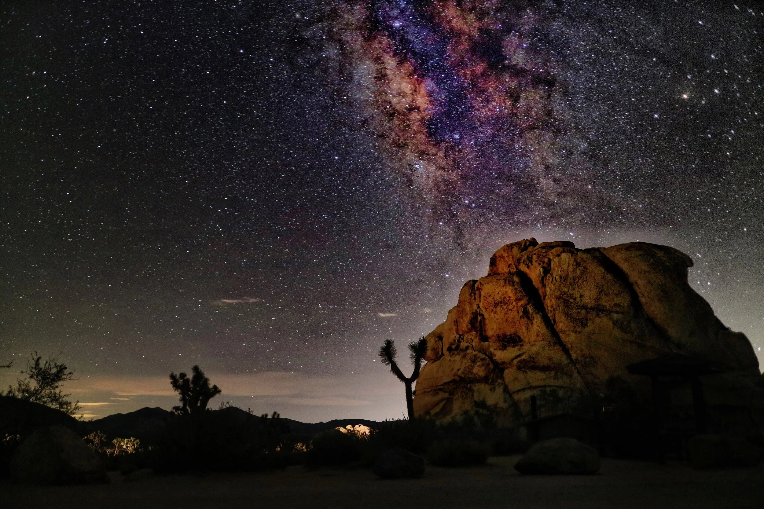 Night sky filled with stars and the Milky Way galaxy above desert landscape with large rock formations, trees, and distant mountains.