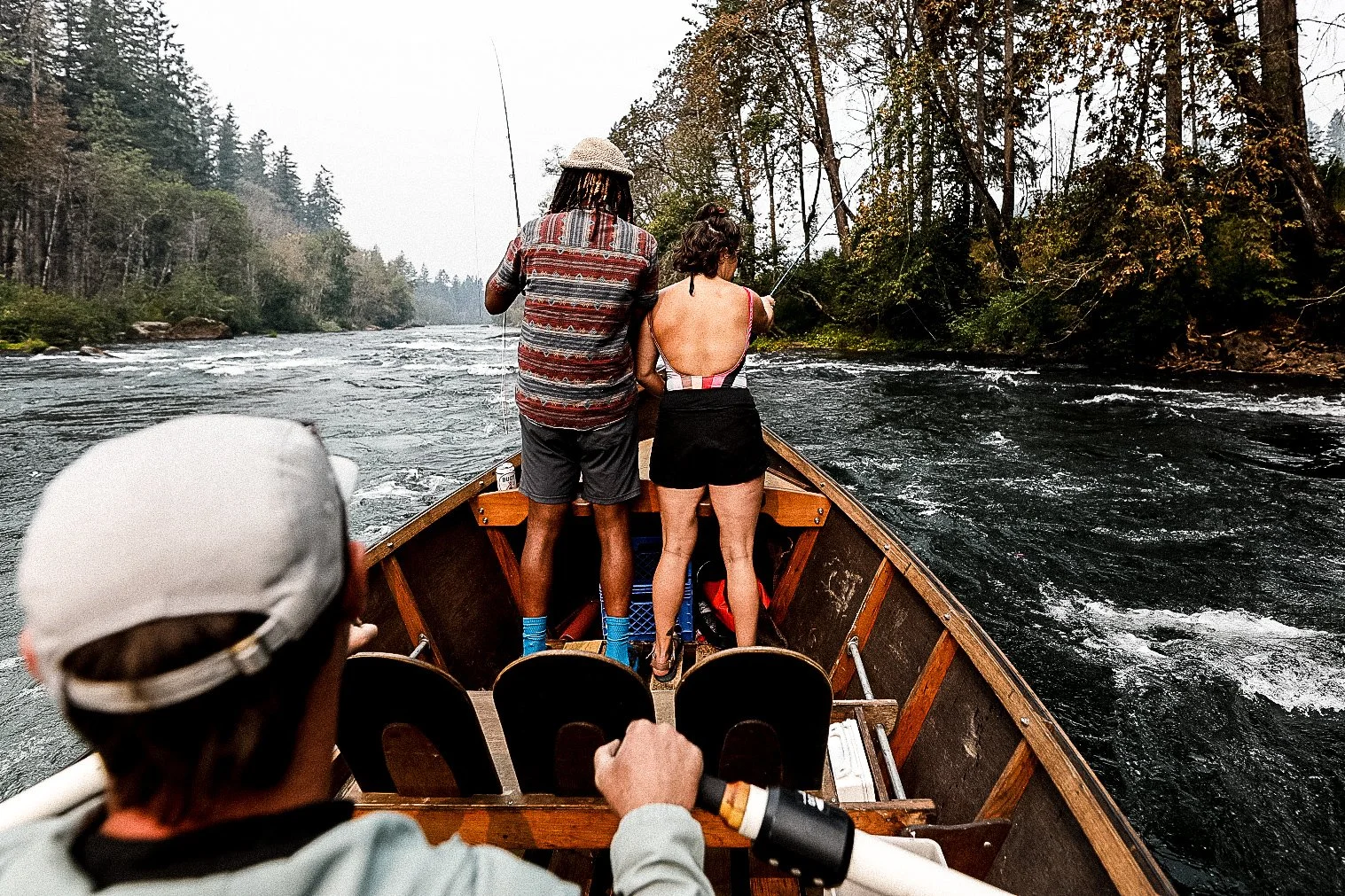 Three people on a boat fishing in a river surrounded by trees.