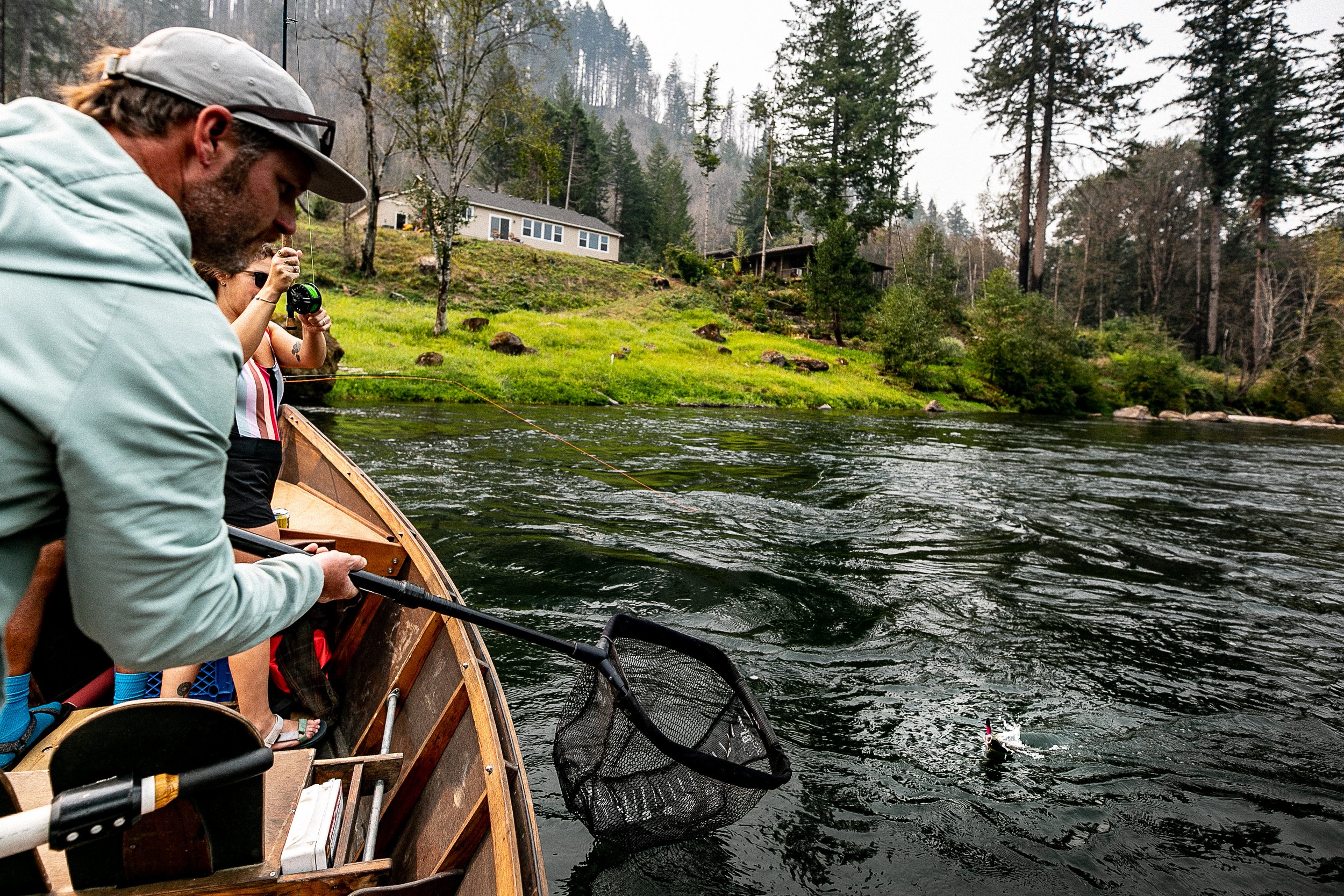 People fishing from a boat on a river with green banks and trees, and houses in the background.