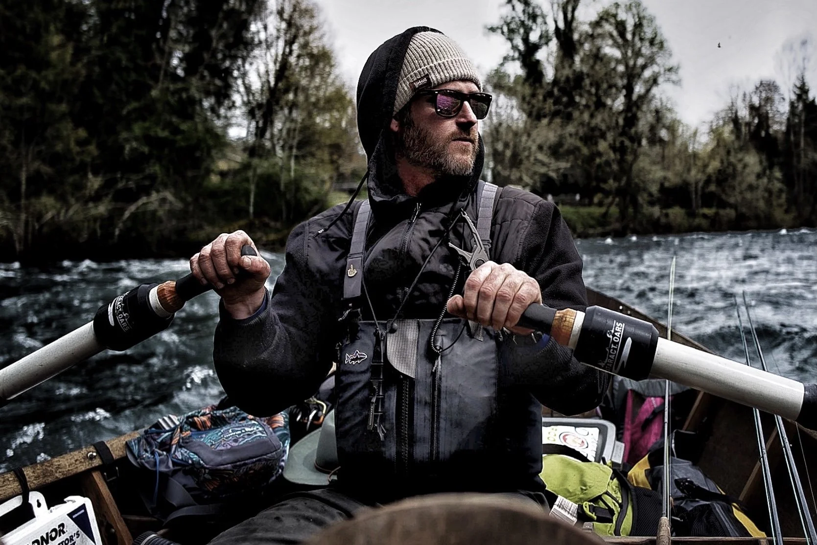 A man with a beard, wearing sunglasses, a beanie, and a black jacket, is rowing a boat on a river with trees in the background.