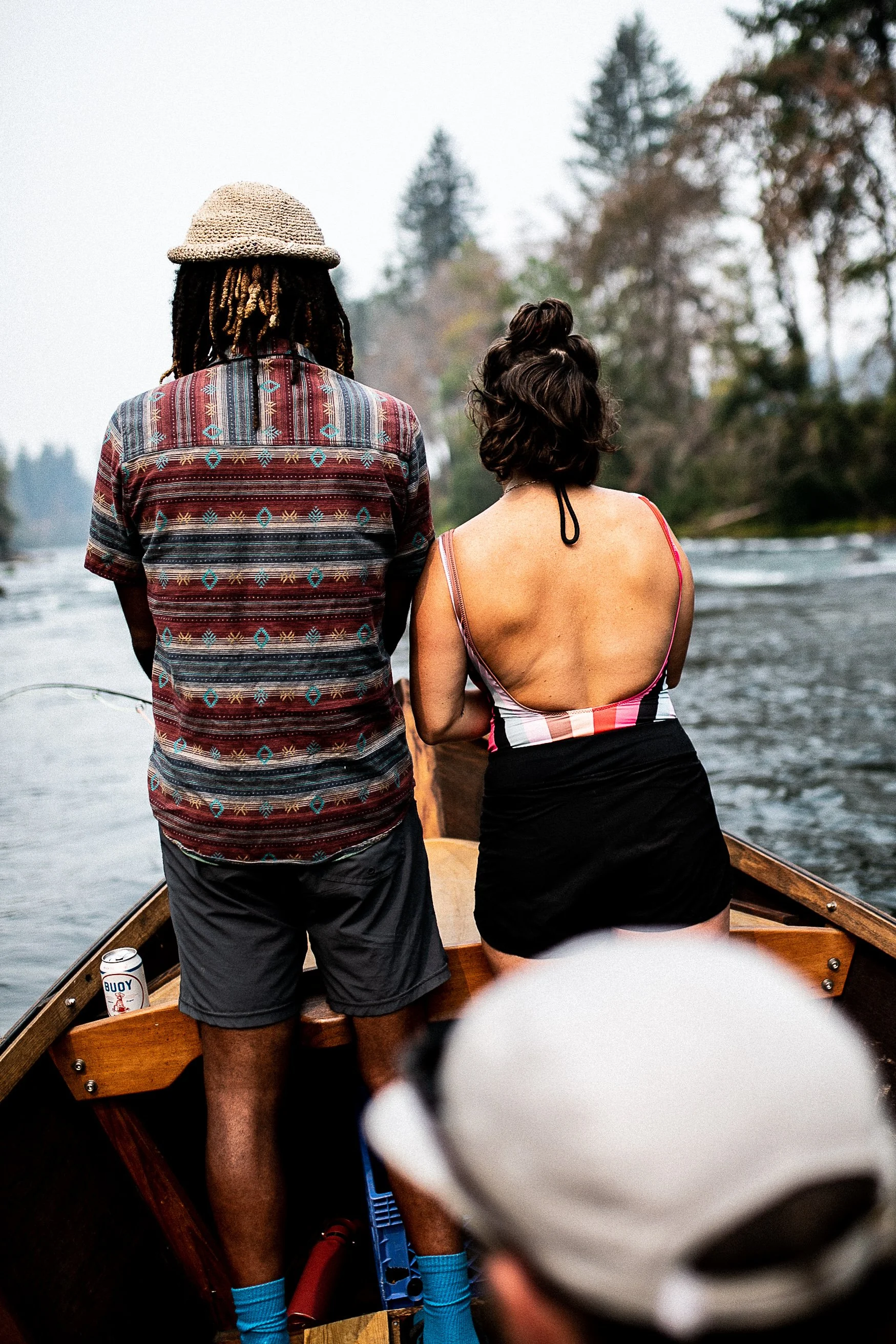 Two people on a wooden boat, with one wearing a patterned shirt and a straw hat, and the other in a colorful dress with a bun hairstyle, heading through a river surrounded by trees.
